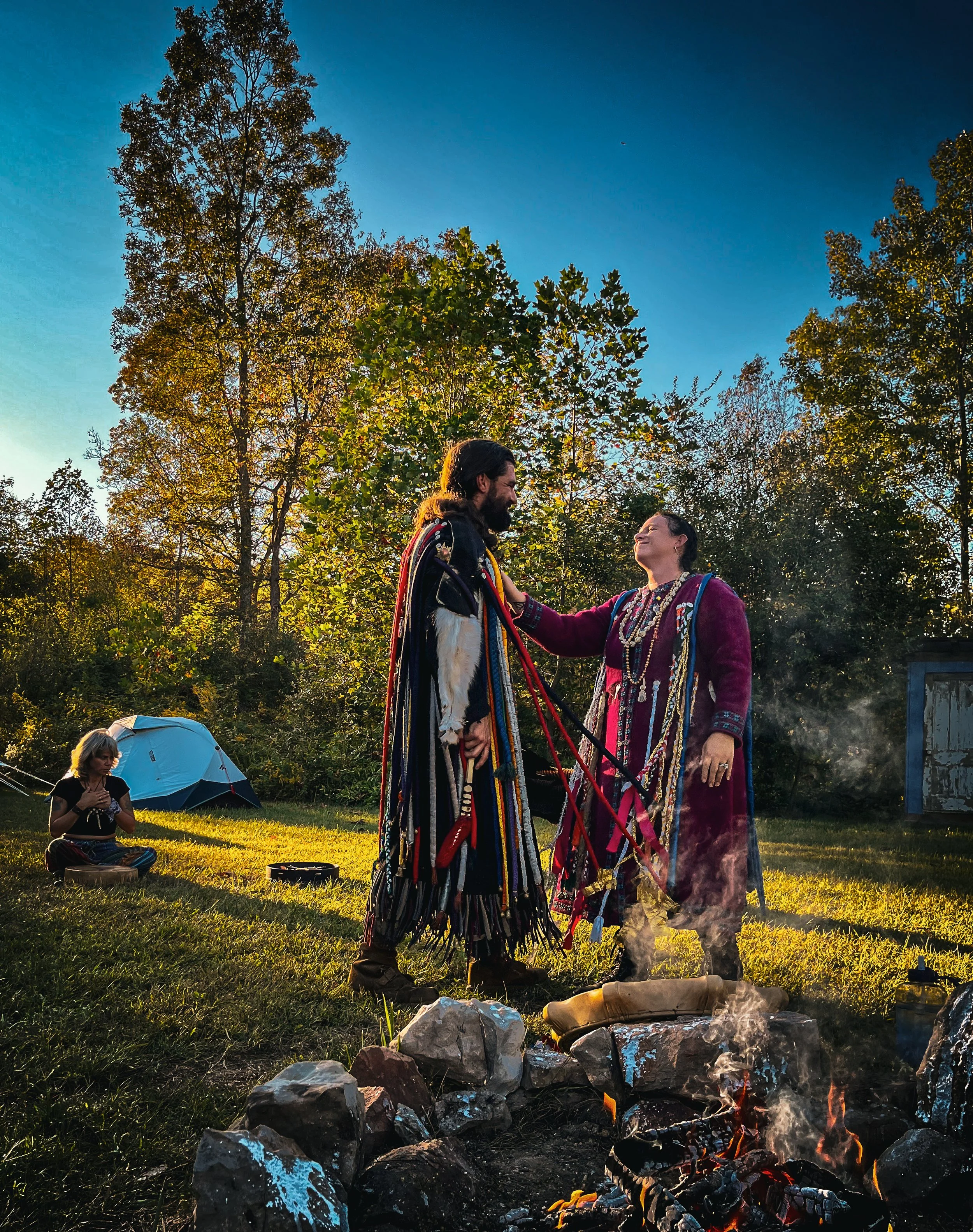 People dressed in traditional clothing performing a ceremonial ritual outdoors during sunset, with one person standing near a campfire and another sitting nearby under an umbrella.