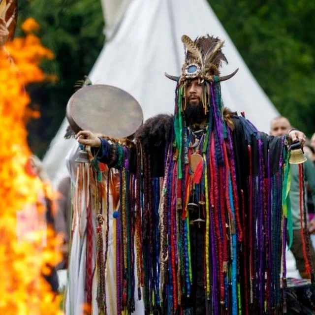 Person wearing an elaborate costume with a feathered headdress, holding a drum and a bell, with a white tipi and orange flames in the background.