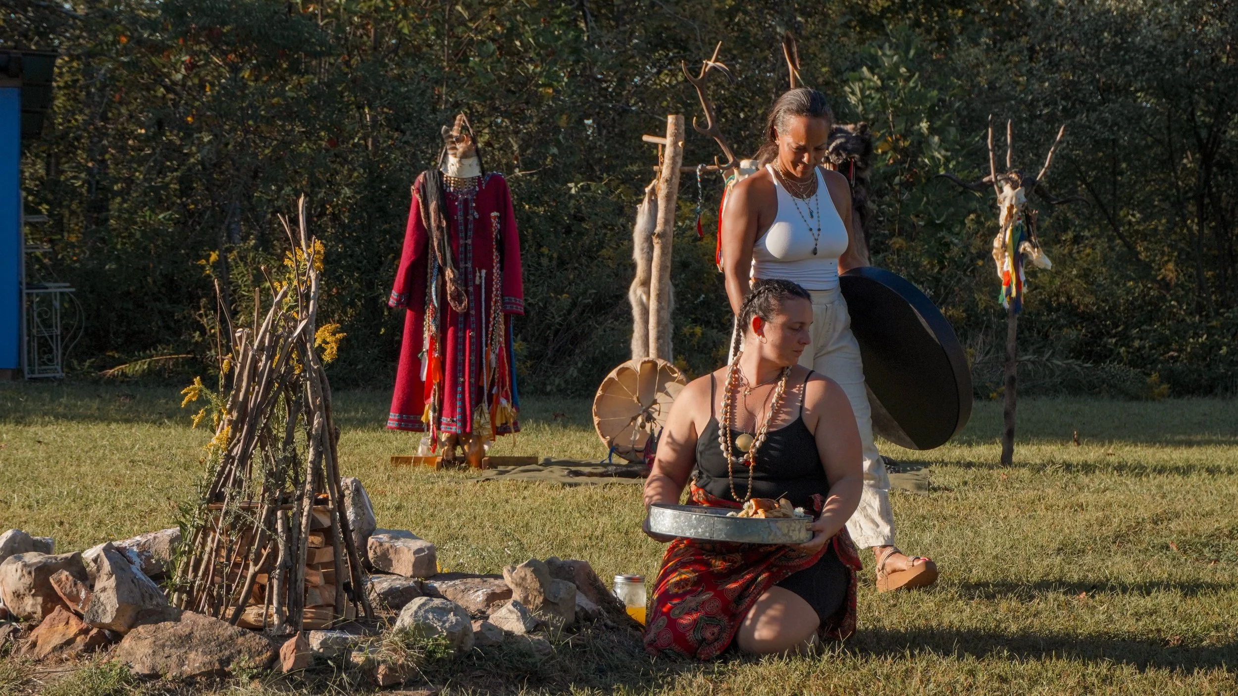 Two women participating in a powwow or Native American cultural ceremony outdoors. One is kneeling with a tray, the other standing with a shield. Traditional regalia, drums, and decorated sticks are visible.