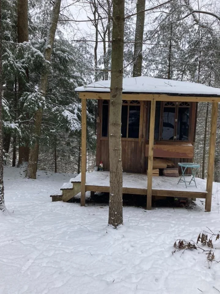 A small wooden cabin on stilts in a snowy forest with trees, featuring a porch with steps, a bench, and a small round table. A place for shamanic and yogic retreats, workshops, and spaces for living altars to humanity and unity with the land.