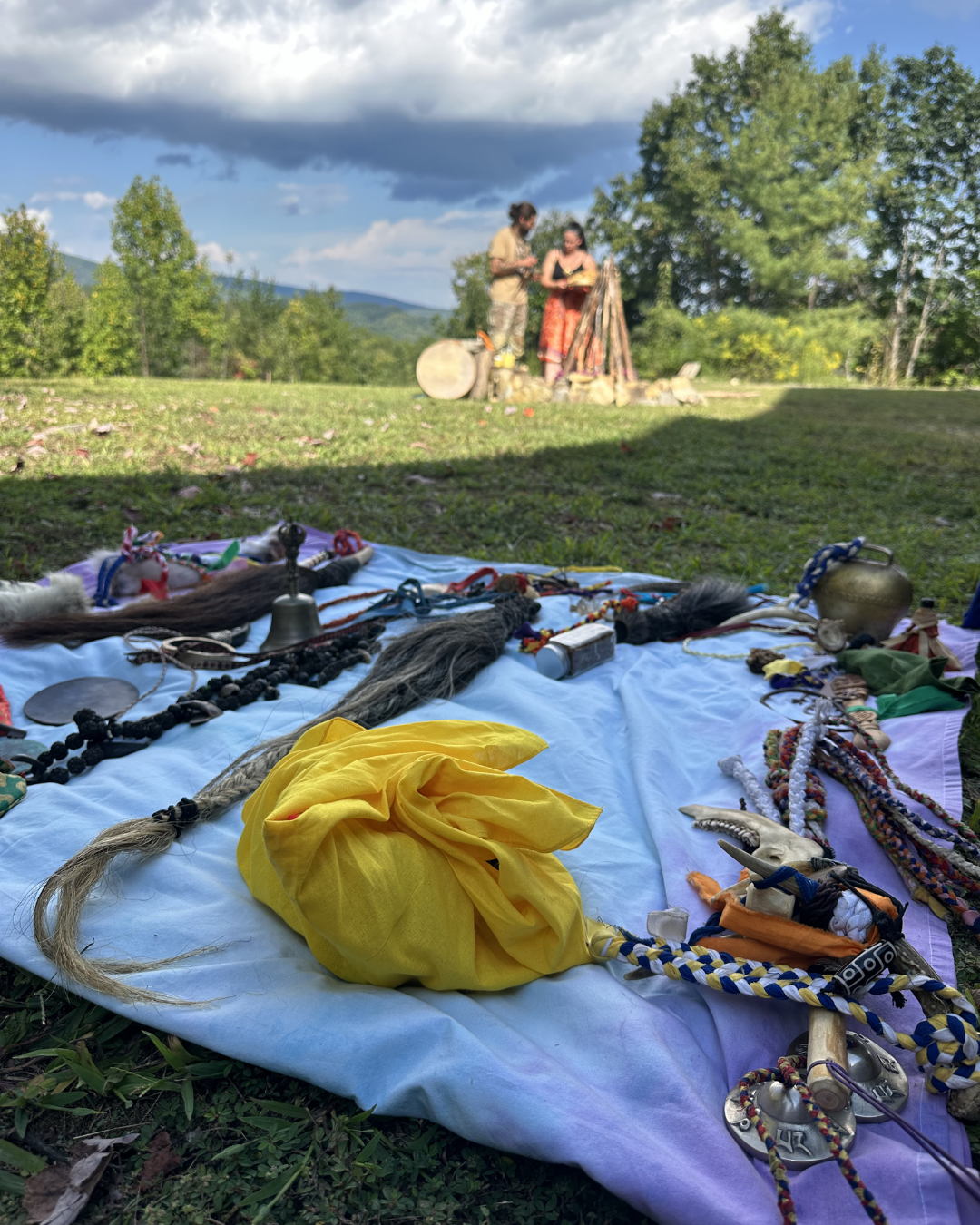 A white blanket on the ground displays various necklaces, cords, and objects, with a yellow cloth in the center. In the background, two women stand near rocks, and a landscape of green trees, mountains, and partly cloudy sky is visible.
