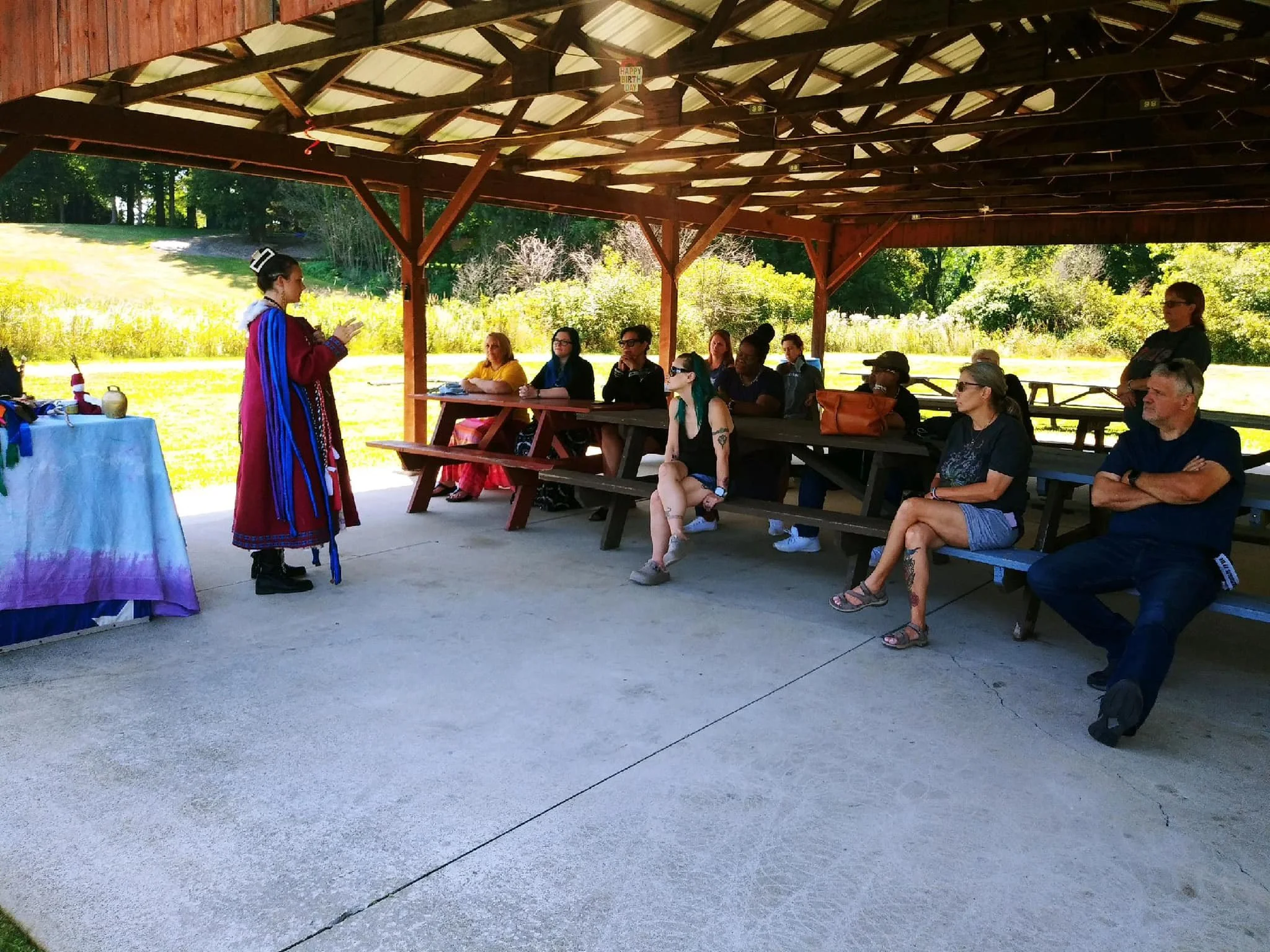 A Native American woman in traditional clothing speaking to an audience at an outdoor pavilion, with seven seated spectators and one standing woman in the background, against a backdrop of greenery and trees.