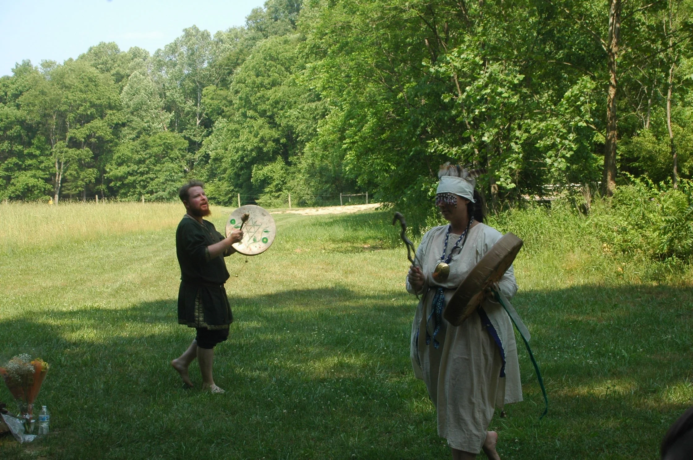 Two people dressed in ancient or fantasy costumes are outdoors in a grassy area with trees. One person, with blonde hair and a blindfold, holds a small staff and a shield. The other, a male with a beard, is holding a drum and a drumstick, appearing t