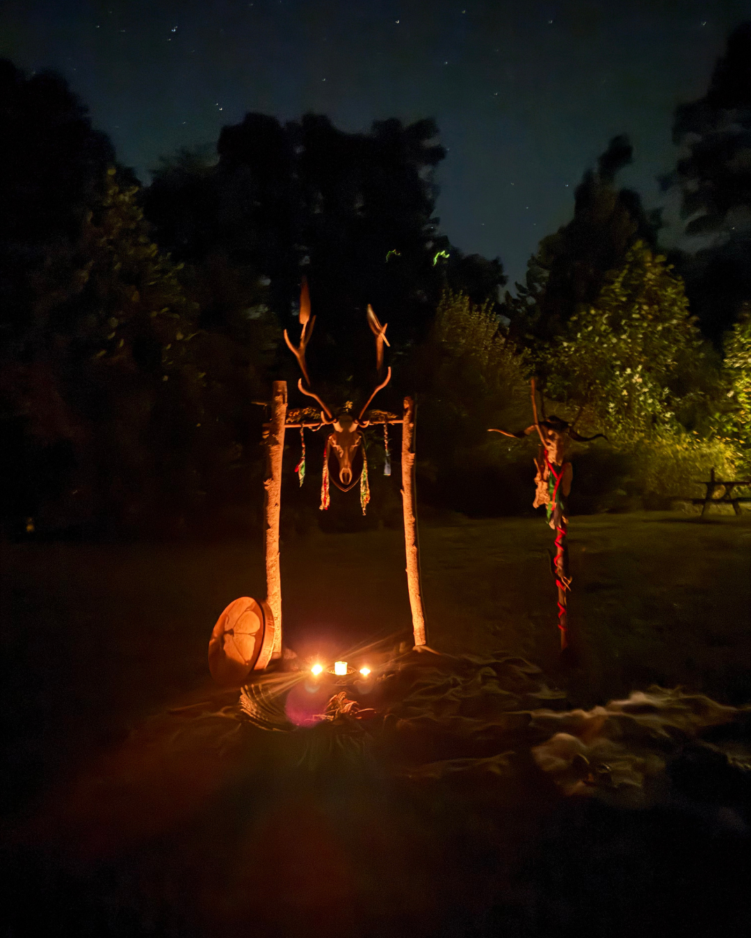 Nighttime outdoor scene with a ceremonial altar decorated with a deer skull with antlers, supported by wooden poles. There is a shield on the ground and candles illuminating the setup, with trees and a starry sky in the background.