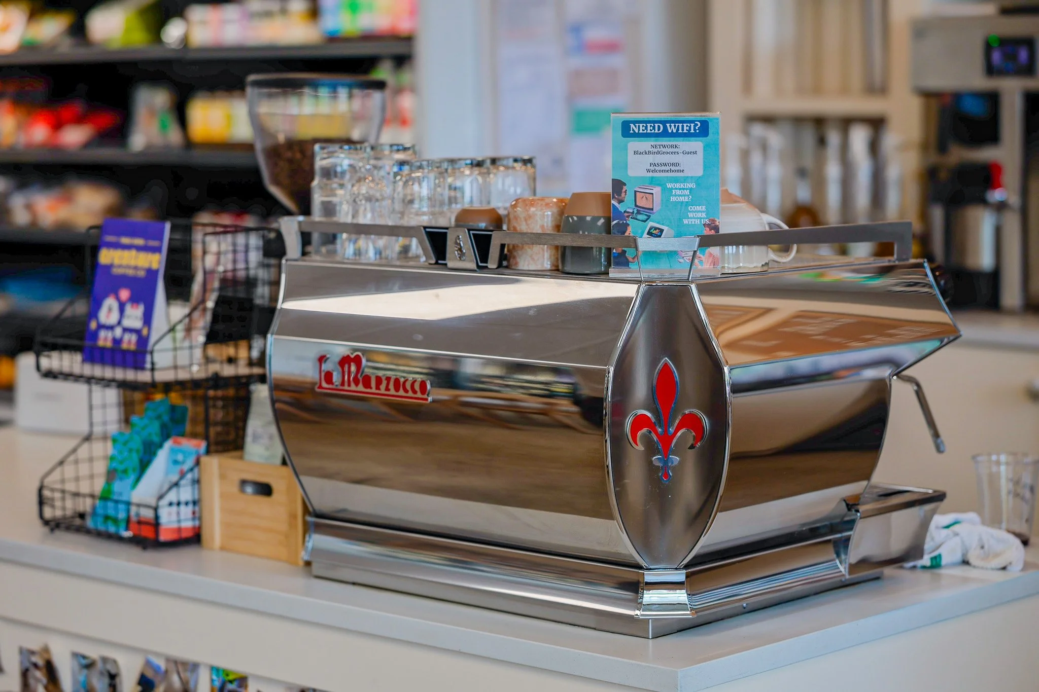 A coffee shop or bakery counter with a shiny, chrome La Marzocco espresso machine on a white countertop, surrounded by small cups, glasses, and a sign about Wi-Fi access.