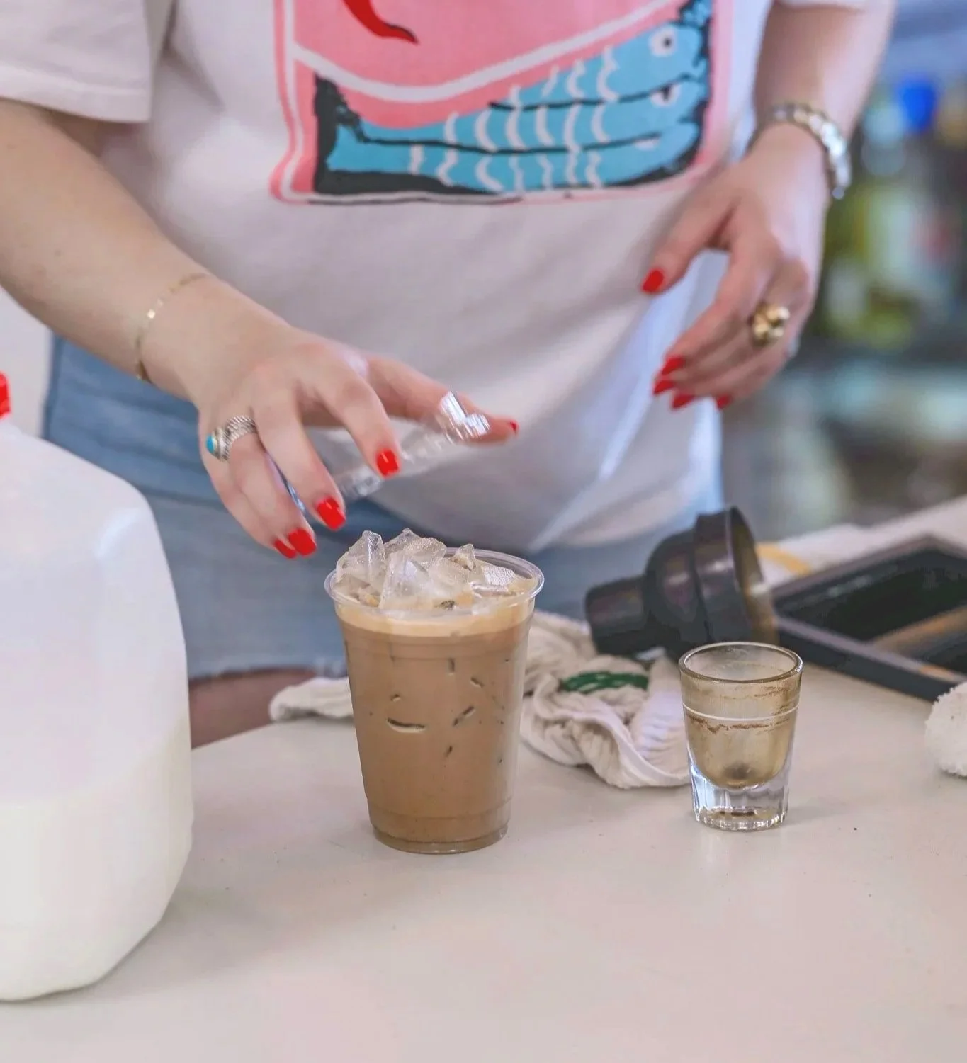 Person with red nails pouring milk into iced coffee at a café counter, surrounded by a glass of water and various items.