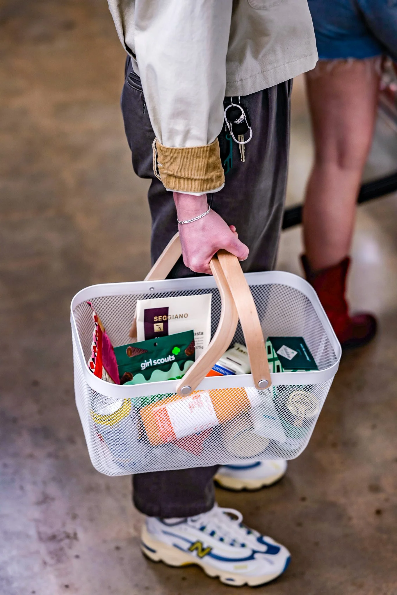 Child holding a white shopping basket filled with various items, wearing a beige jacket with brown sleeve cuffs, dark pants, and white sneakers with blue and yellow accents, standing indoors on a concrete floor.
