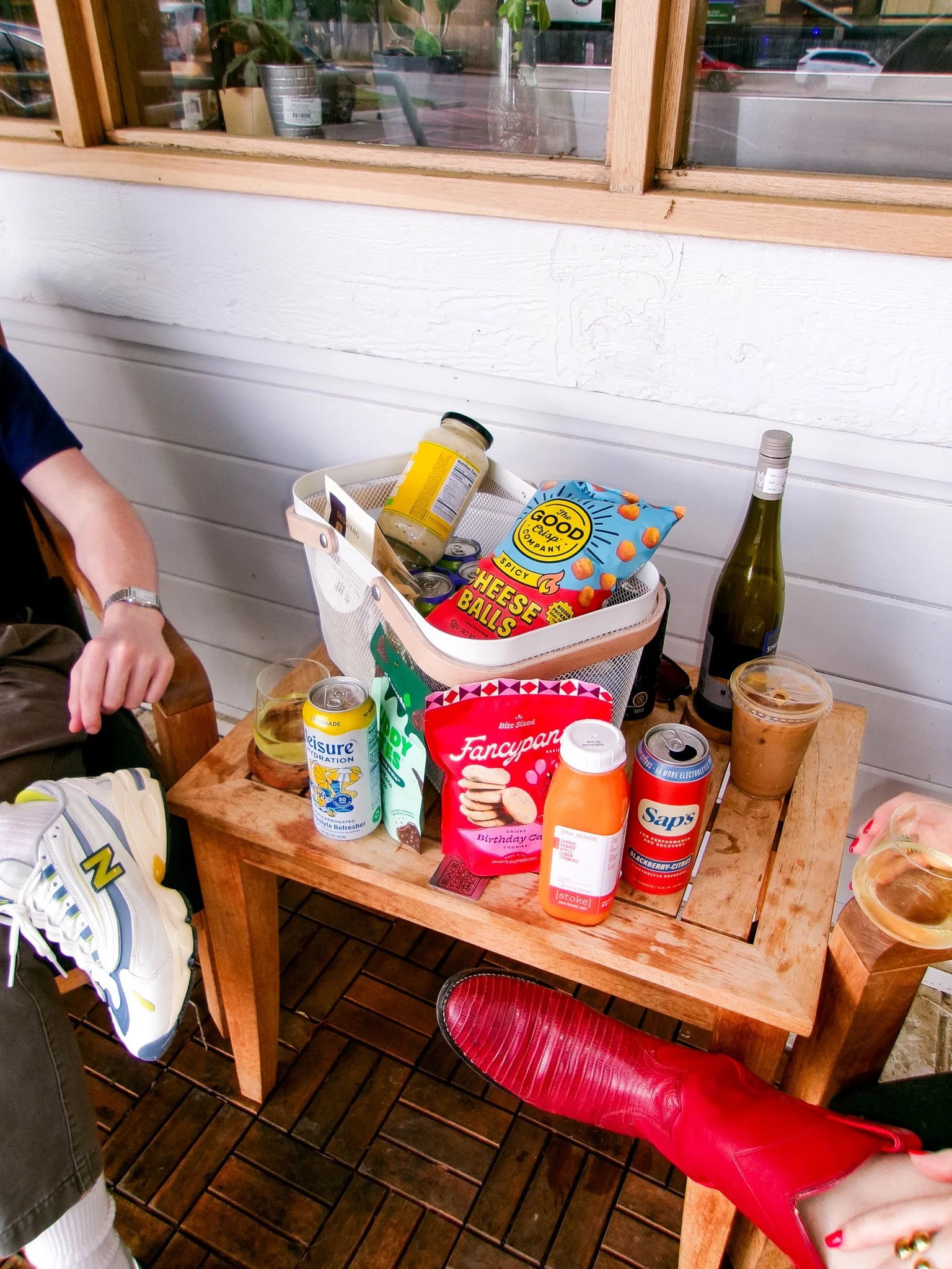 A wooden table with various snacks and drinks on it, including a bag of cheese balls, cans of beverages, a bottle of white wine, and a glass of iced coffee, with a pair of red rain boots visible at the bottom right corner.