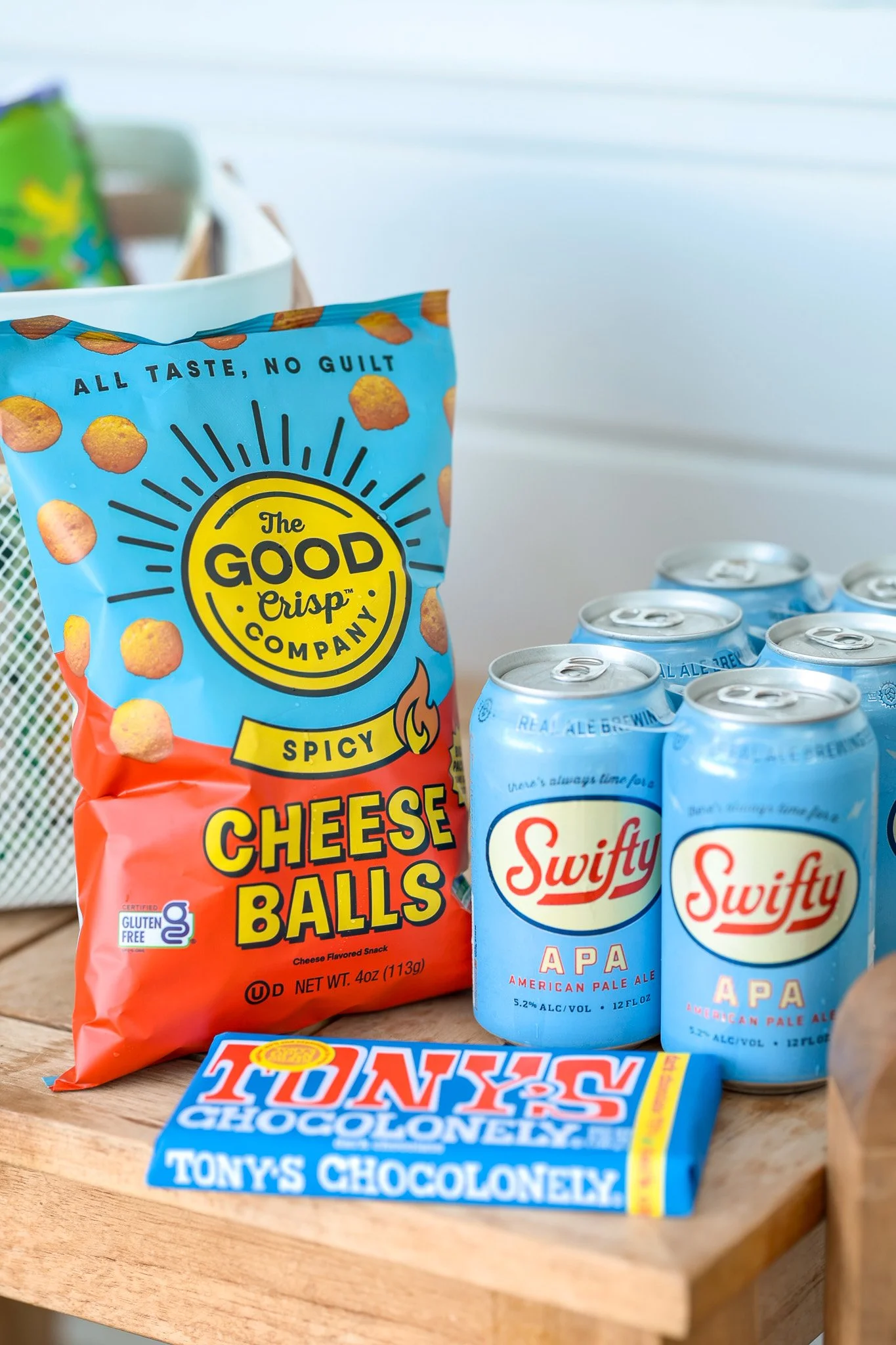 A bag of spicy cheese balls, six cans of Swifty APA American Pale Ale beer, and a bar of Tony's Chocolonely chocolate on a wooden surface.