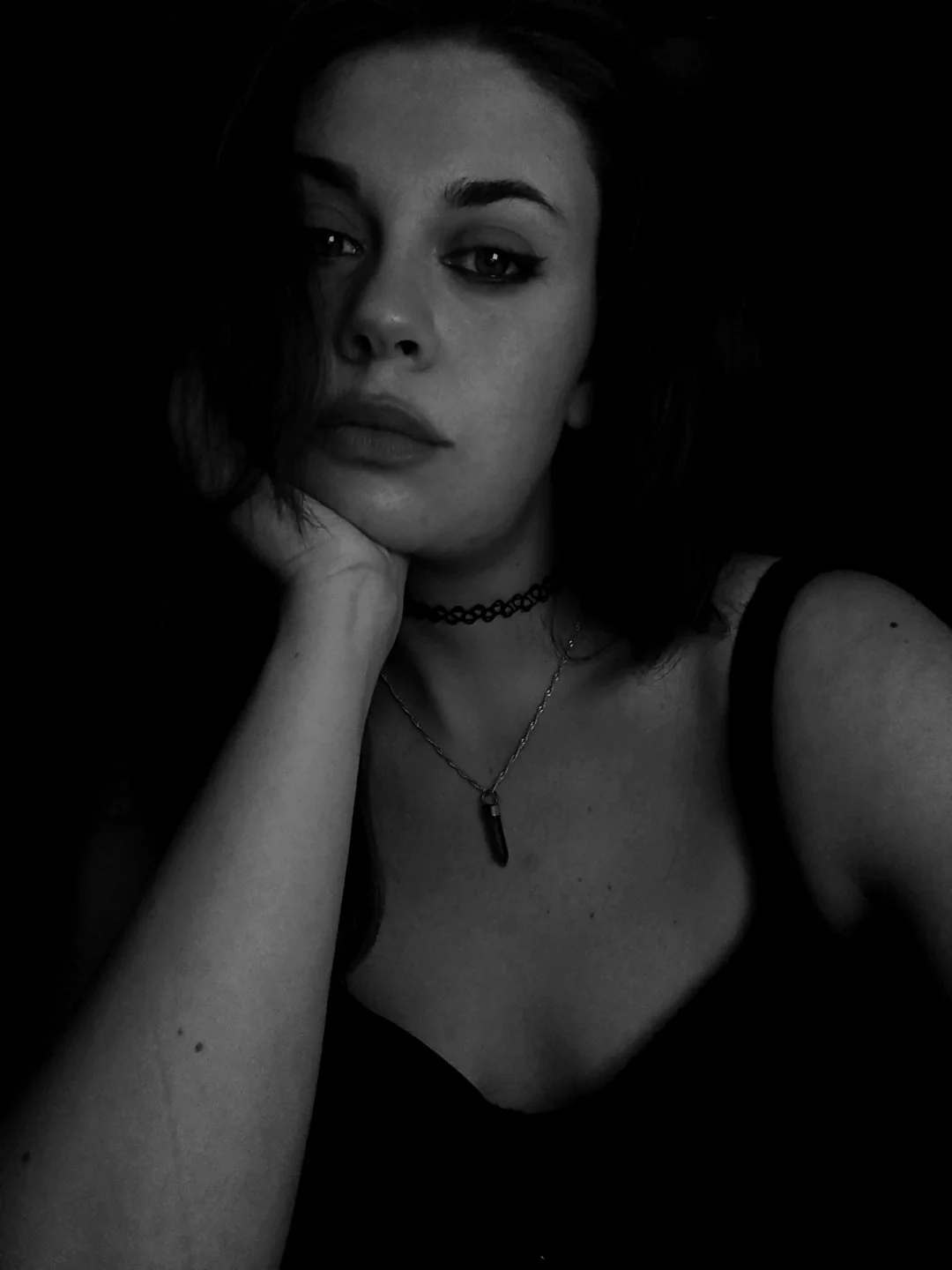 Black and white portrait of a woman with dark hair, wearing a choker and necklace, resting her chin on her hand, against a dark background.