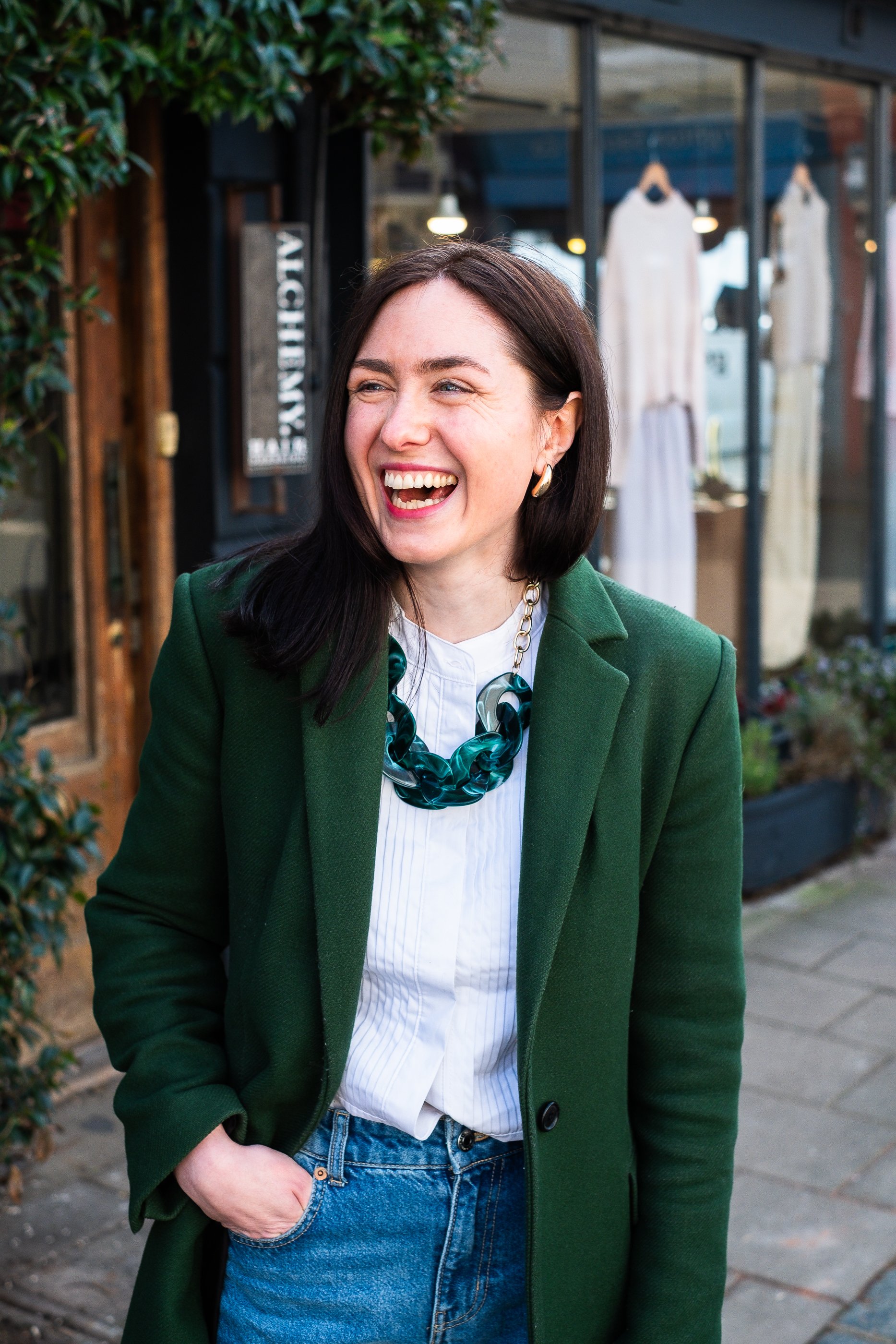 A white woman with dark brown shoulder length hair, wearing a dark green coat, white shirt and dark green chunky necklace  laughing to the right