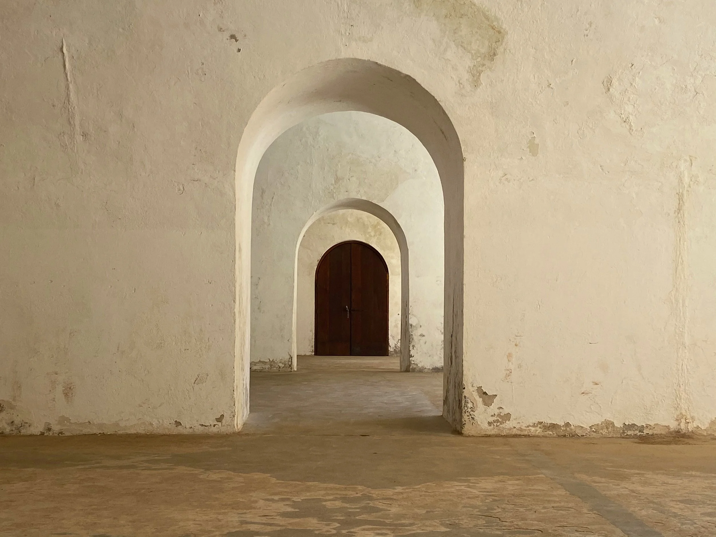 A series of white, worn, arched doorways or windows leading to a brown wooden door in the distance, set in a textured, weathered beige wall.