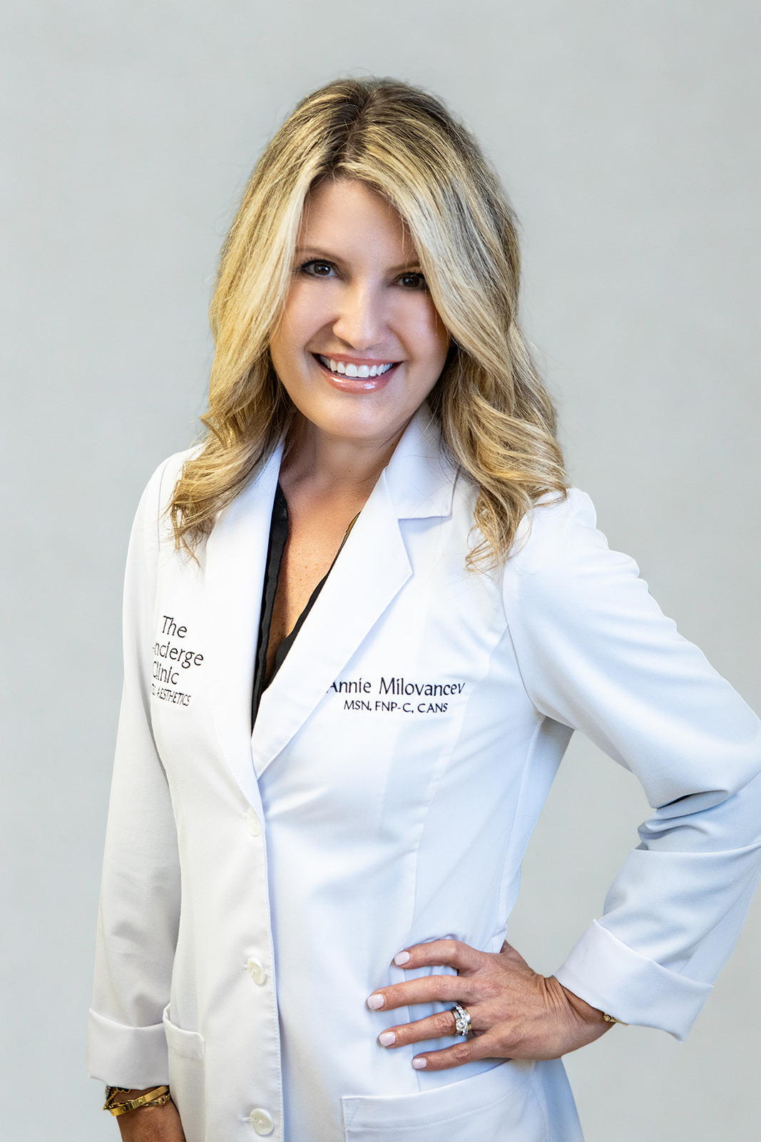A smiling woman with blonde hair wearing a white coat with text, posing with her hand on her hip against a plain background.