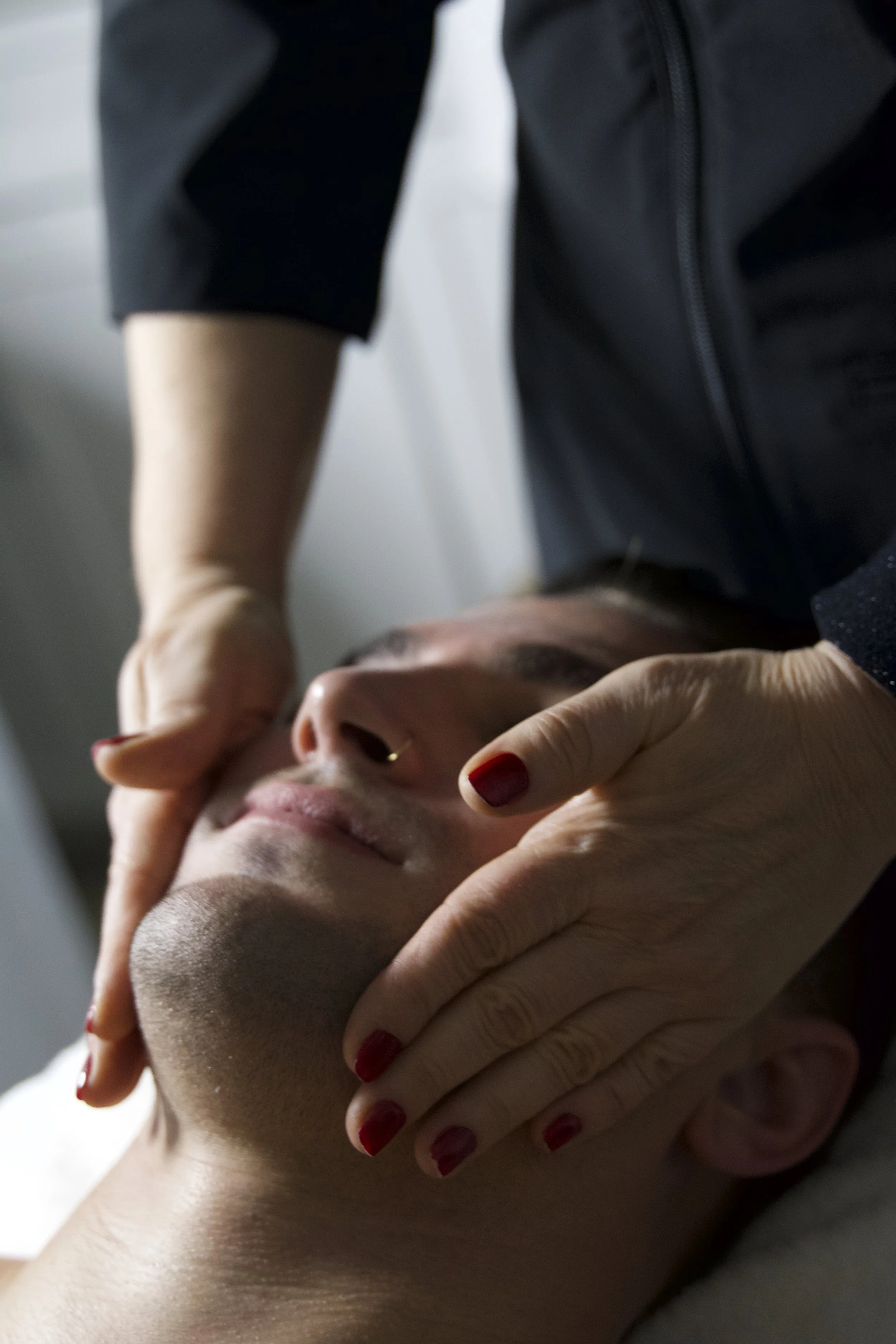 A person receiving a facial massage with their eyes closed, lying down. Hands are gently massaging the face, and the person has red nail polish.