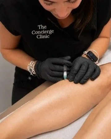 A medical professional in black gloves and black scrubs labeled 'The Concierge Clinic' preparing to give an injection into a person's arm.