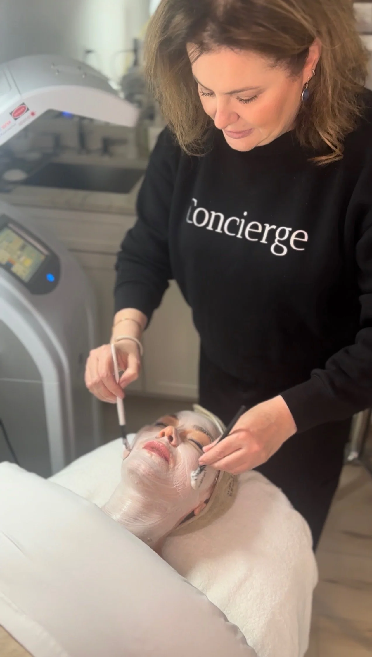 A woman receiving a facial treatment with a facial mask applied, lying on a spa bed while a professional performs skincare procedures.