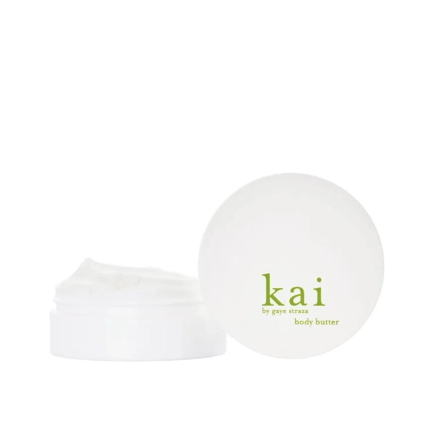 A white round container labeled "kai" by Gaye Straza, body butter, with the lid next to it and some of the body butter visible inside.