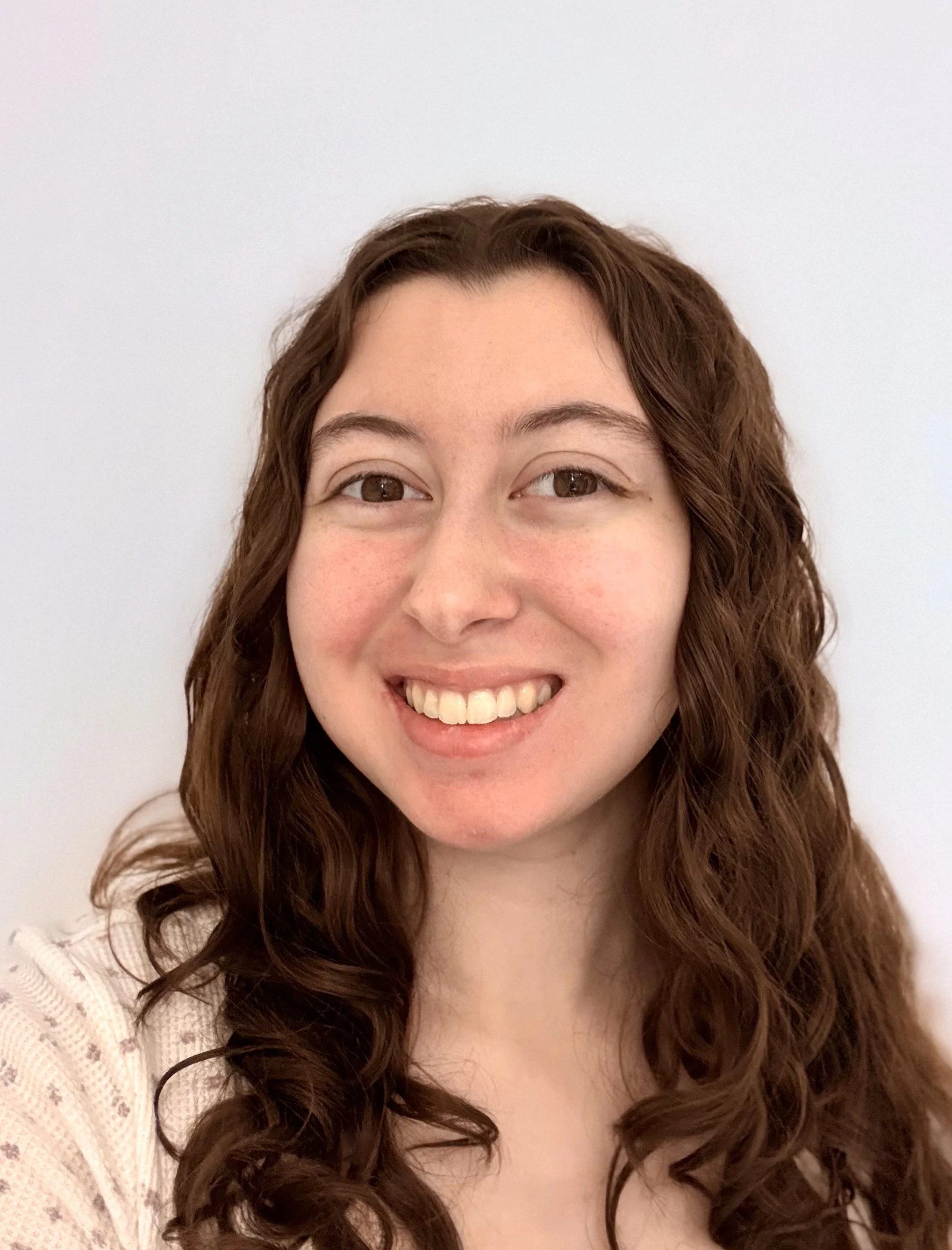 A young woman with curly brown hair smiling in front of a plain white background.