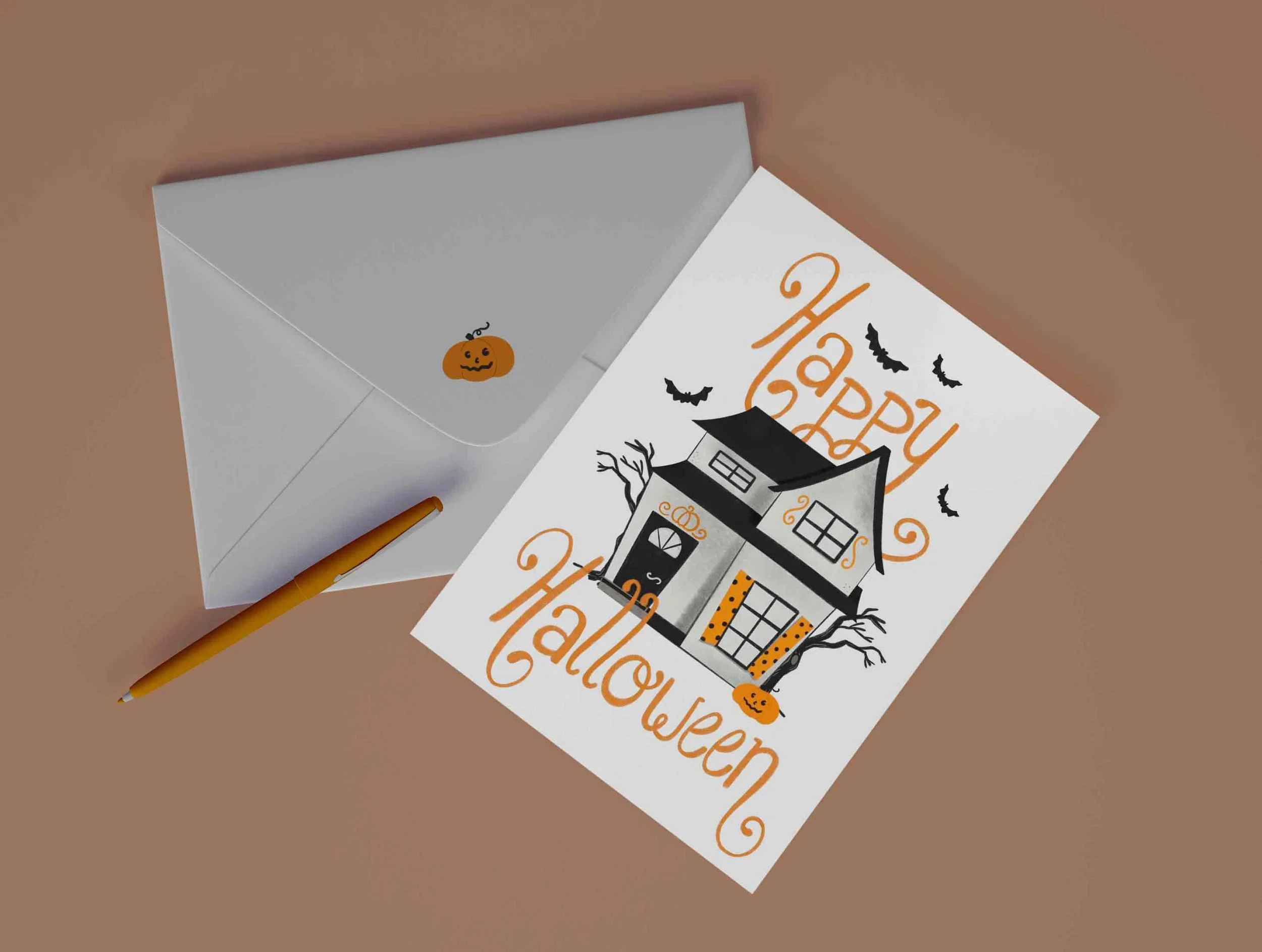 A Halloween greeting card with a haunted house illustration, bats, and pumpkins, beside a white envelope and an orange pen on a brown surface.