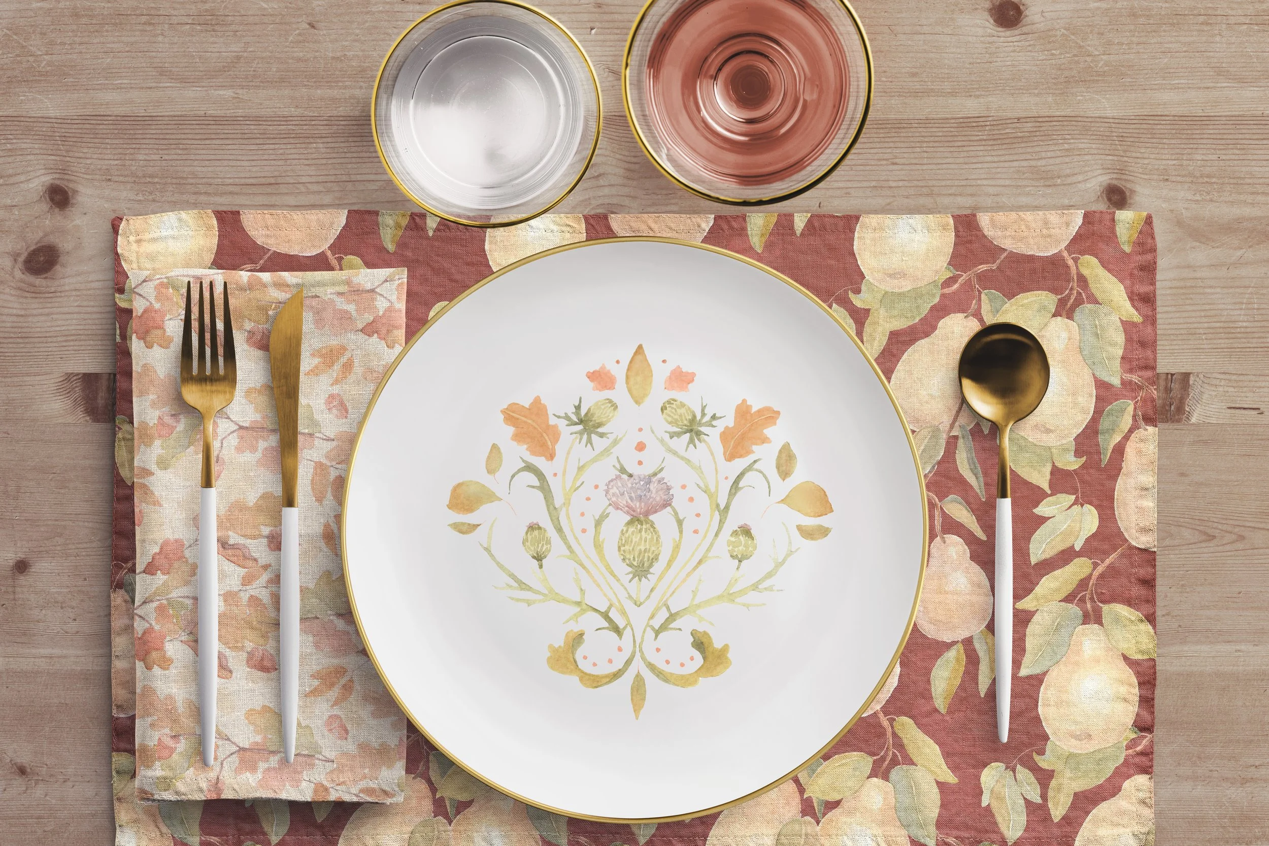 Elegant table setting featuring a floral-printed plate with gold edges, gold and white cutlery, a gold spoon, a napkin with a floral pattern, and two glasses of water and rosé wine on a wooden table with a floral tablecloth.