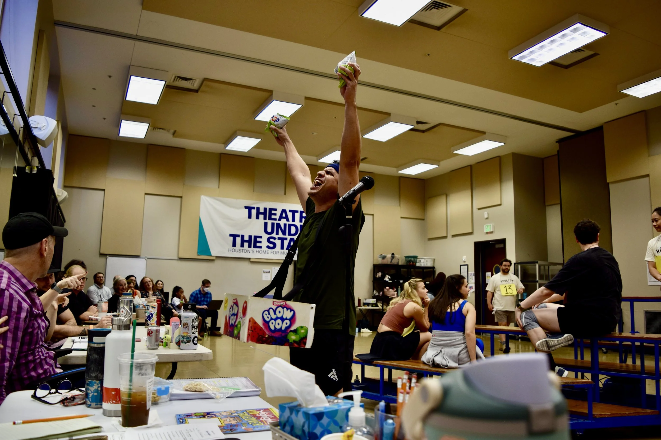 A person celebrating with arms raised in a room with a crowd, tables, and snacks, during a theater or performance event.