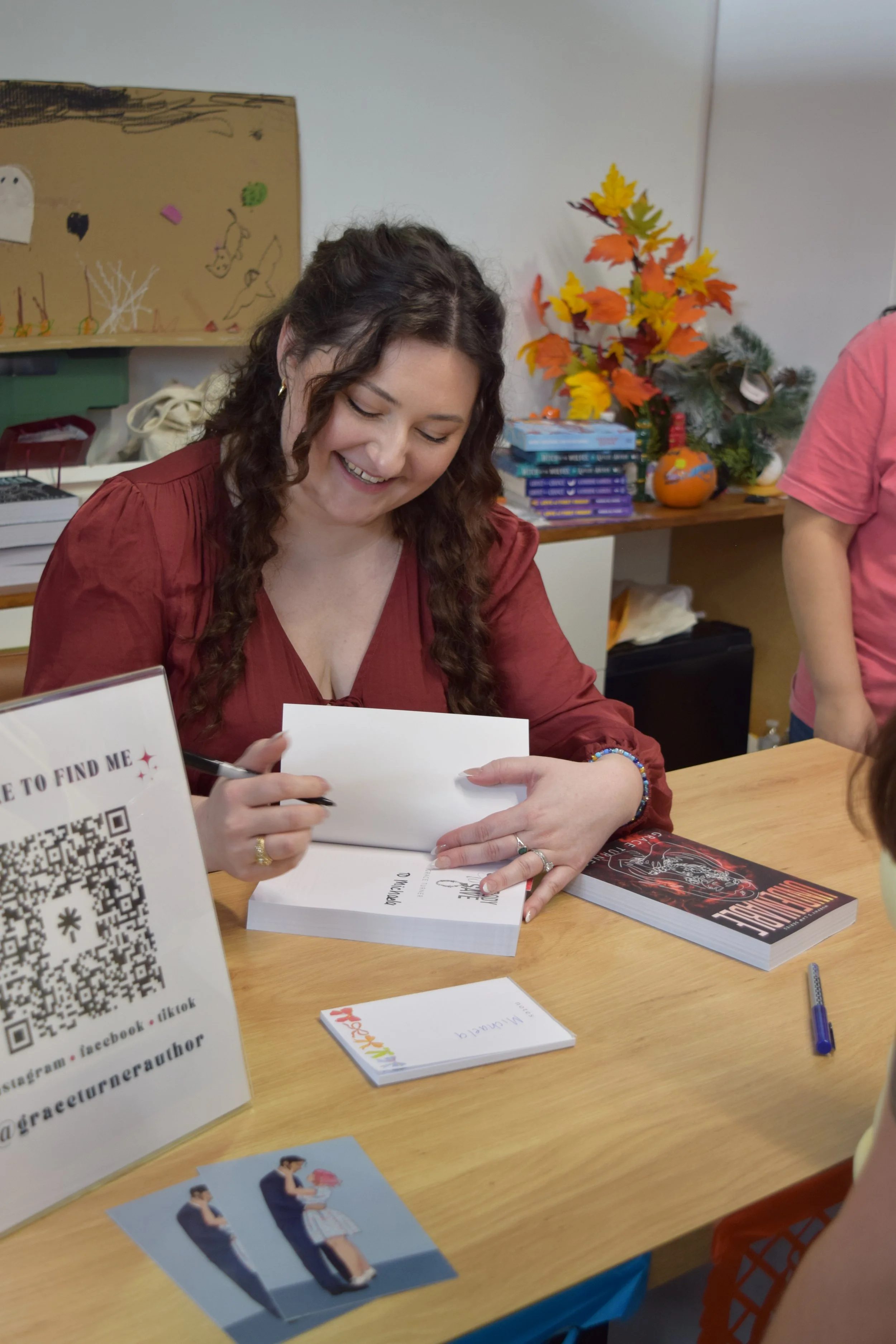 A woman with long curly hair, wearing a red blouse, is sitting at a table signing a book. There are books and flyers on the table, and a sign with a QR code and social media information. In the background, there are autumn-themed decorations and a st