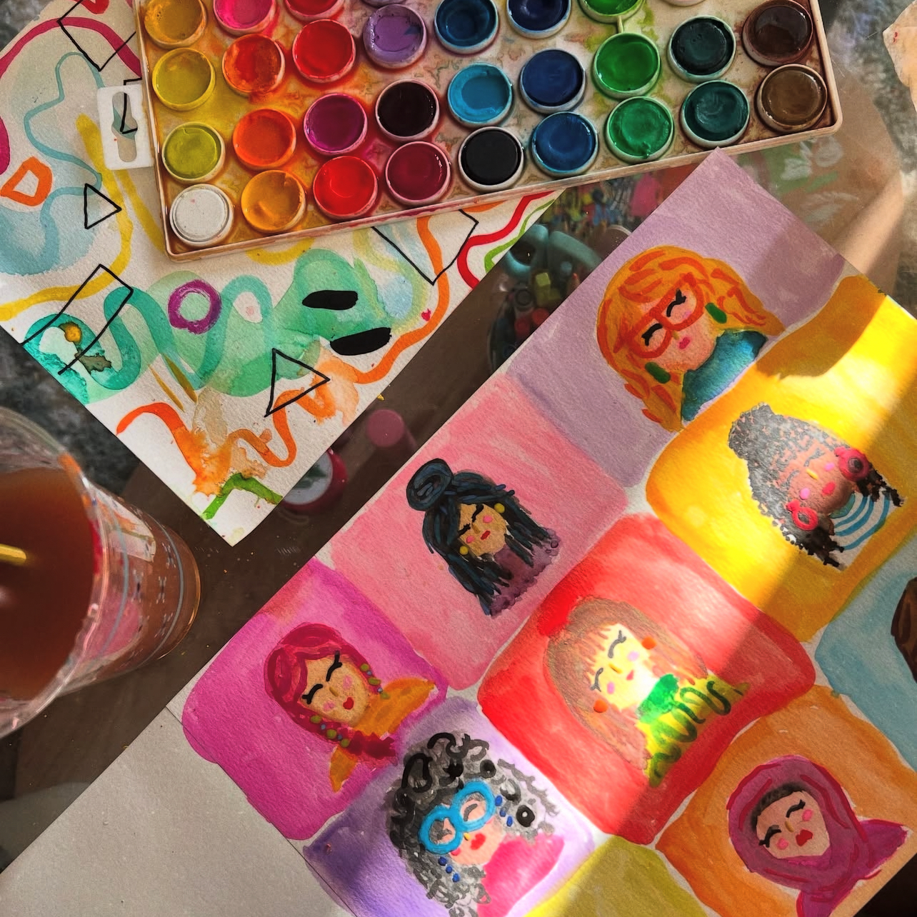 A collection of watercolor paintings of diverse women, each with different hairstyles and accessories, alongside a watercolor palette with various colors and a glass of iced tea.