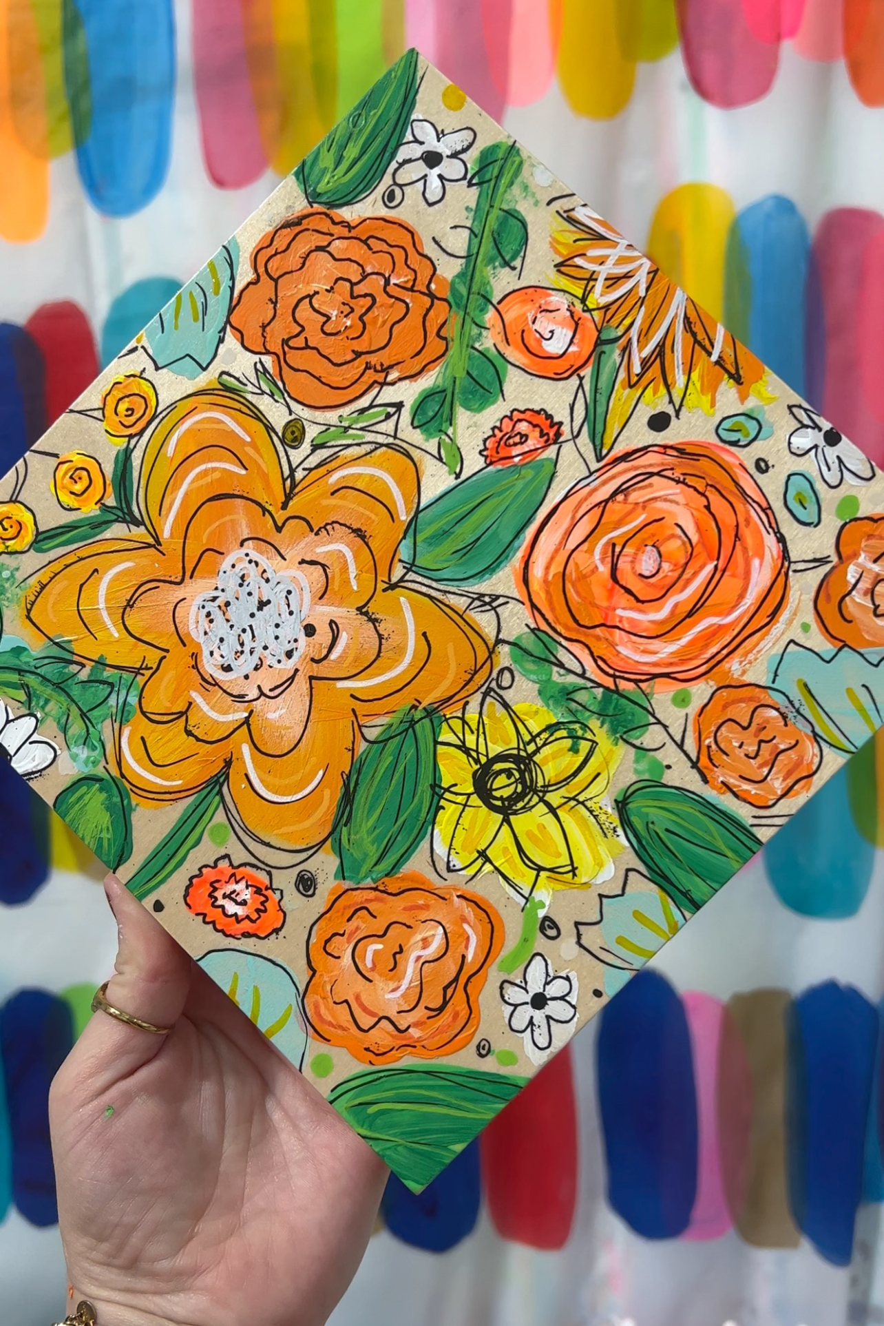 A hand holding a colorful piece of artwork with floral patterns, including orange, yellow, and white flowers, and green leaves, with a background of rainbow-colored vertical stripes.