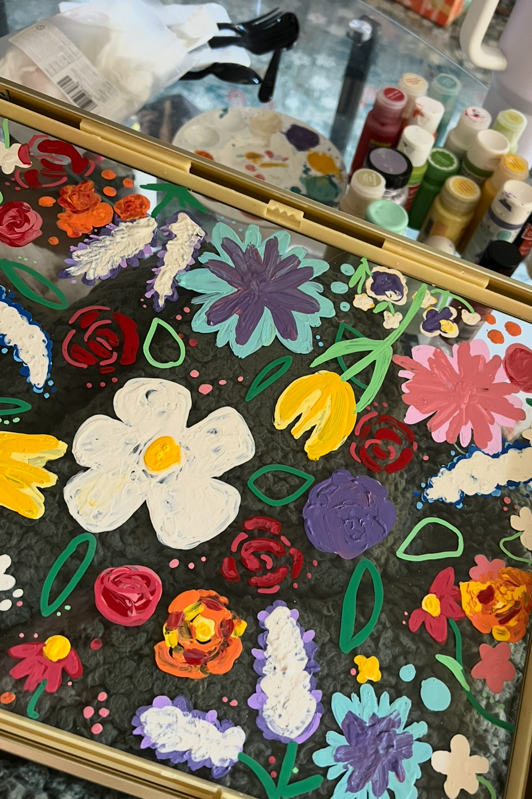 Colorful floral painting on glass, featuring various flowers painted in bright colors including white, purple, yellow, pink, red, orange, and lavender, with green leaves and stems, surrounded by bottles of paint and painting supplies.