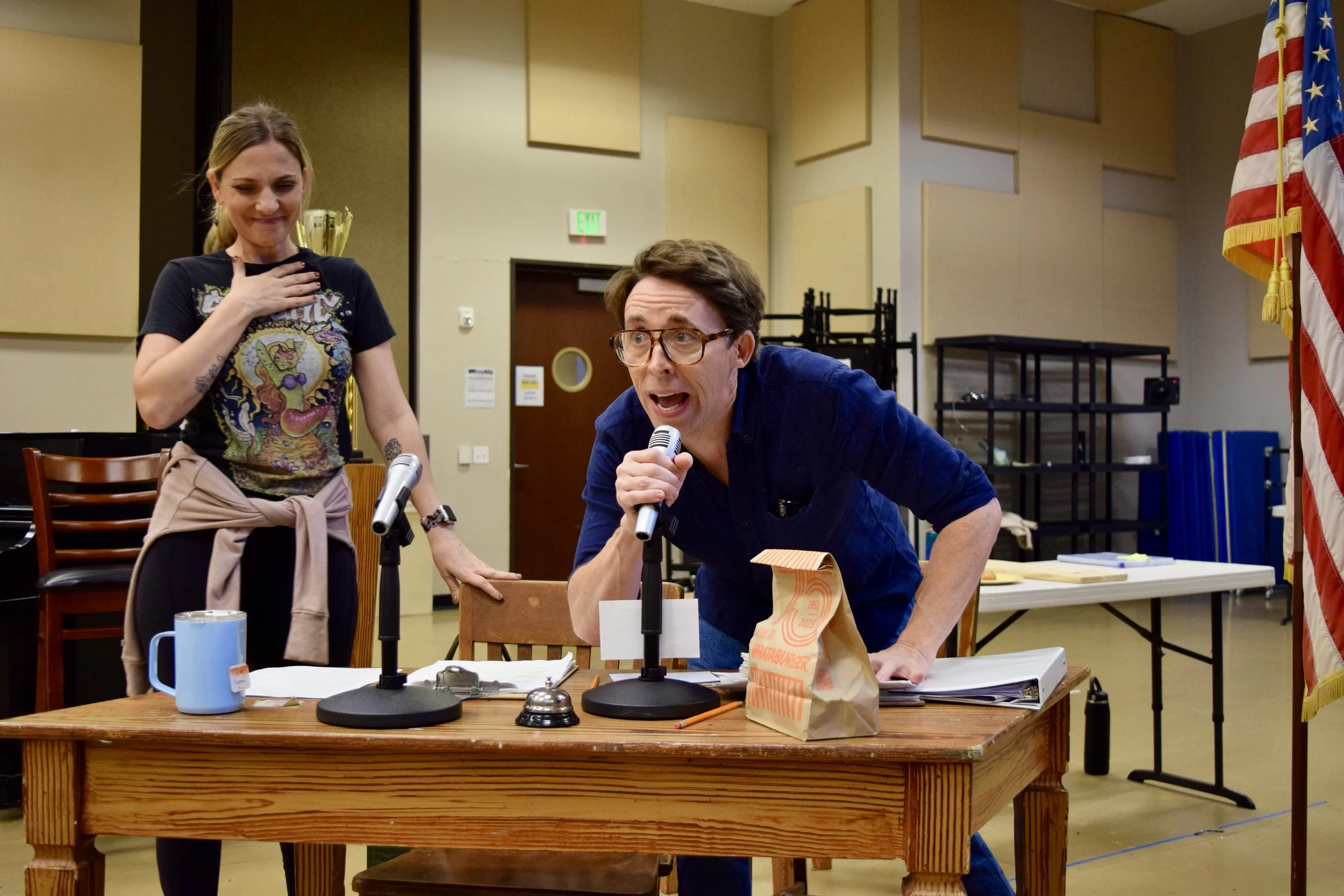 A man and a woman are in a room, likely a community center or classroom, with the man speaking passionately into a microphone at a wooden table. The woman stands beside him, smiling with her hand over her chest, wearing a graphic t-shirt and bib over