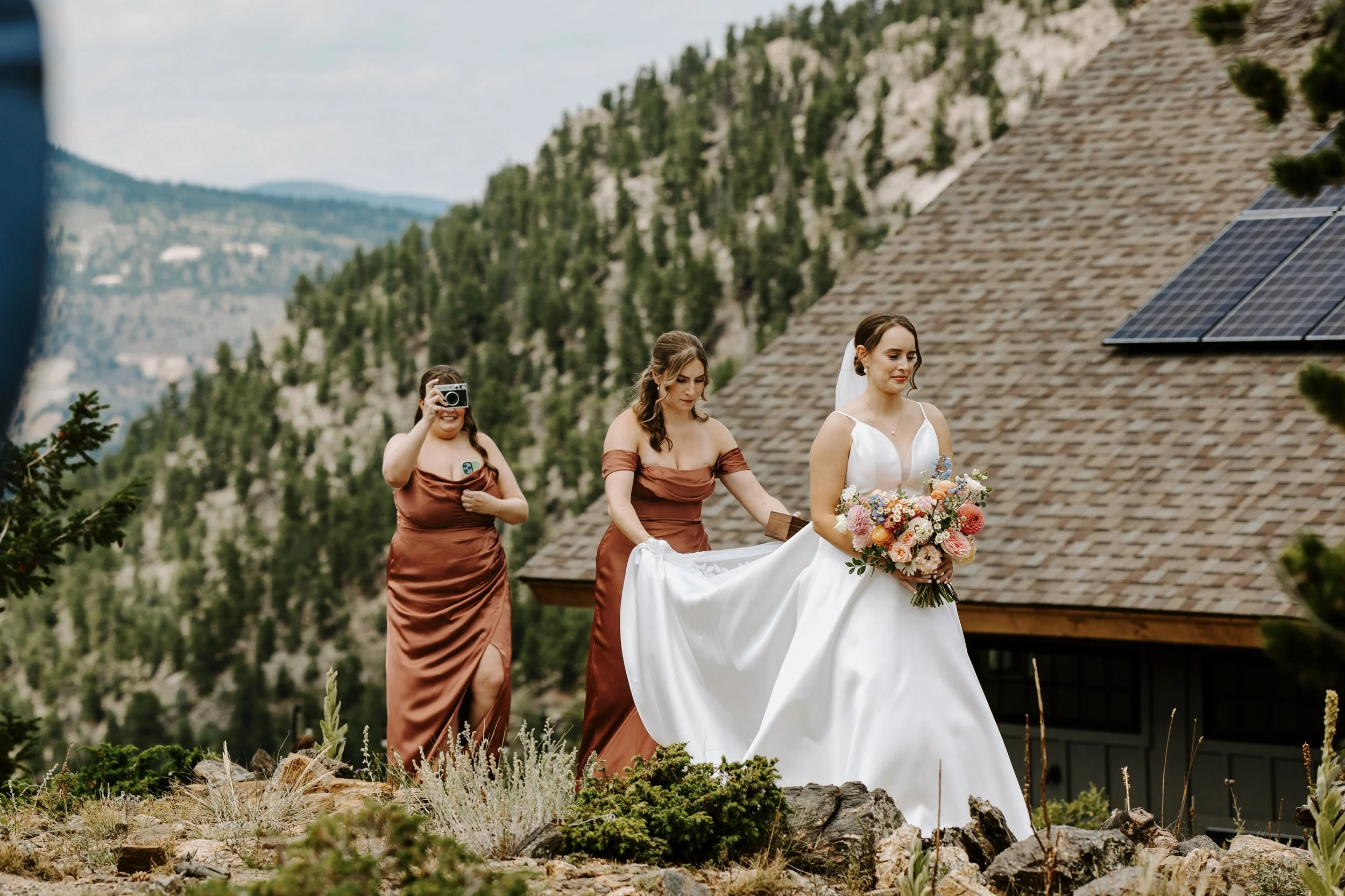 A bride in a white wedding gown holding a bouquet of colorful flowers walking outdoors, accompanied by two bridesmaids in copper-colored dresses, one of whom is taking a photo with a vintage camera, with a mountain landscape and a house with solar panels in the background.
