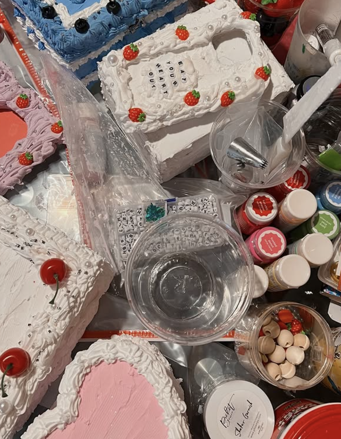 Assorted cakes decorated with strawberries, icing, and pearls, surrounded by cake decorating supplies such as sprinkles, icing, and cake toppers.