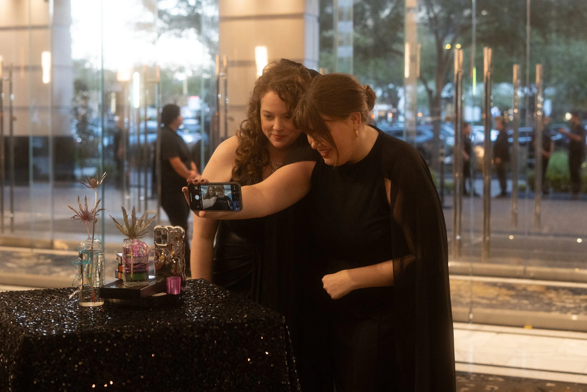 Two women in black dresses taking a selfie at an event with glass doors and trees in the background.