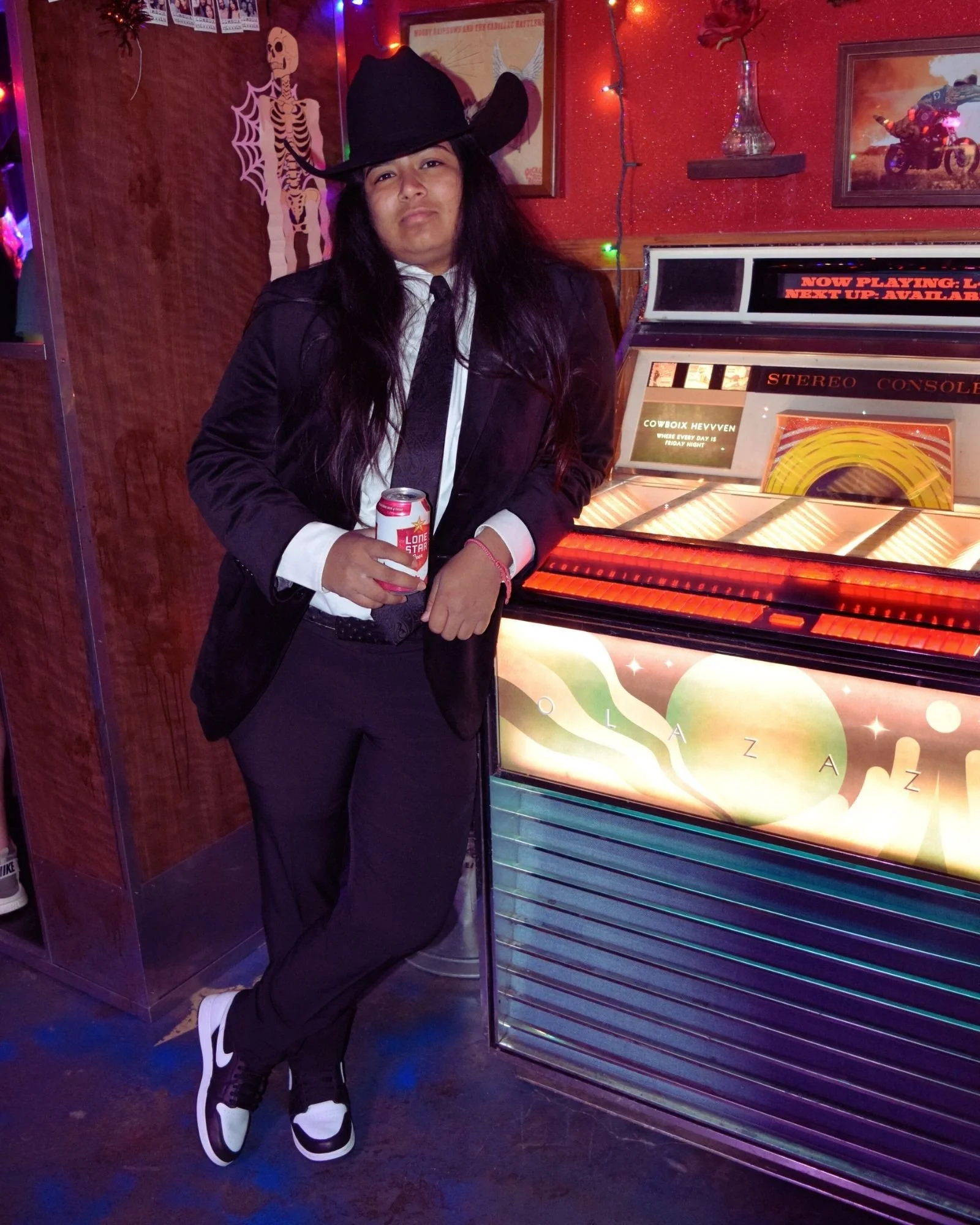 Person dressed as a cowboy, wearing a black hat, black suit, white shirt, and black tie, standing in front of a vintage jukebox in a dimly lit room with colorful lights and decor.