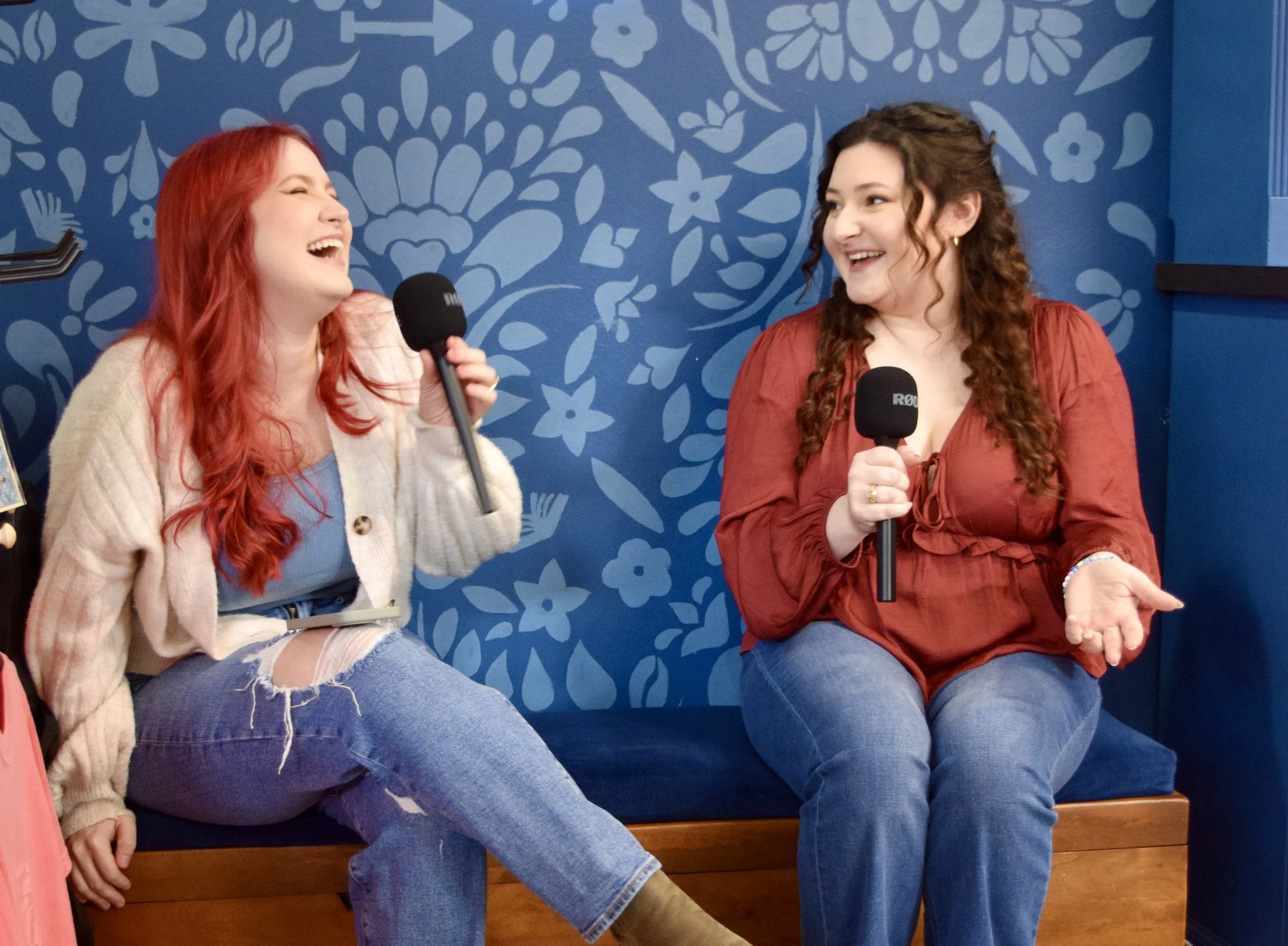 Two women sitting on a bench holding microphones, laughing and talking during an interview in a room with blue floral wallpaper.