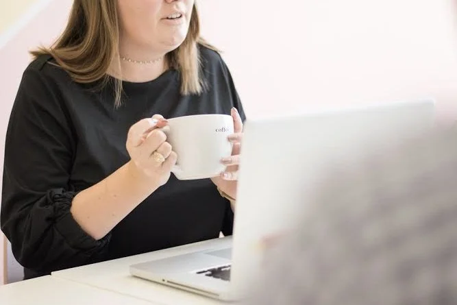 A woman sitting at a desk holding a white mug near a laptop, with a light-colored wall in the background.