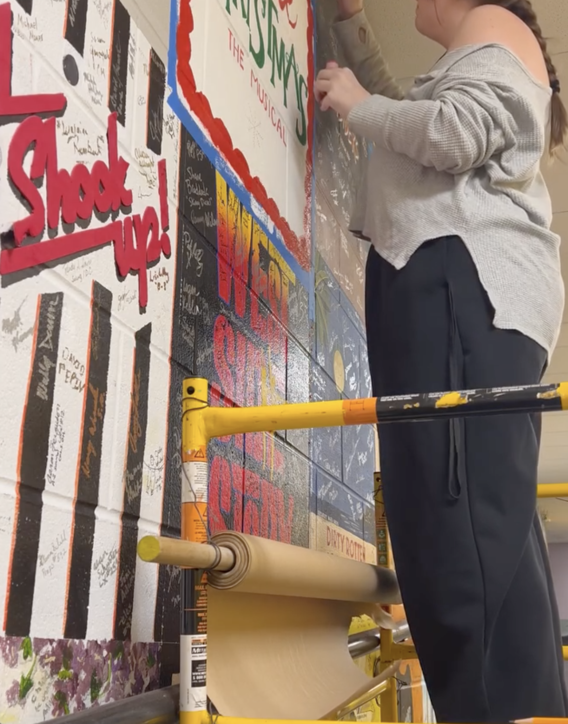 Person signing a large poster board covered with messages and drawings, with the prominent words "Shout Up!" and references to music and community activism.
