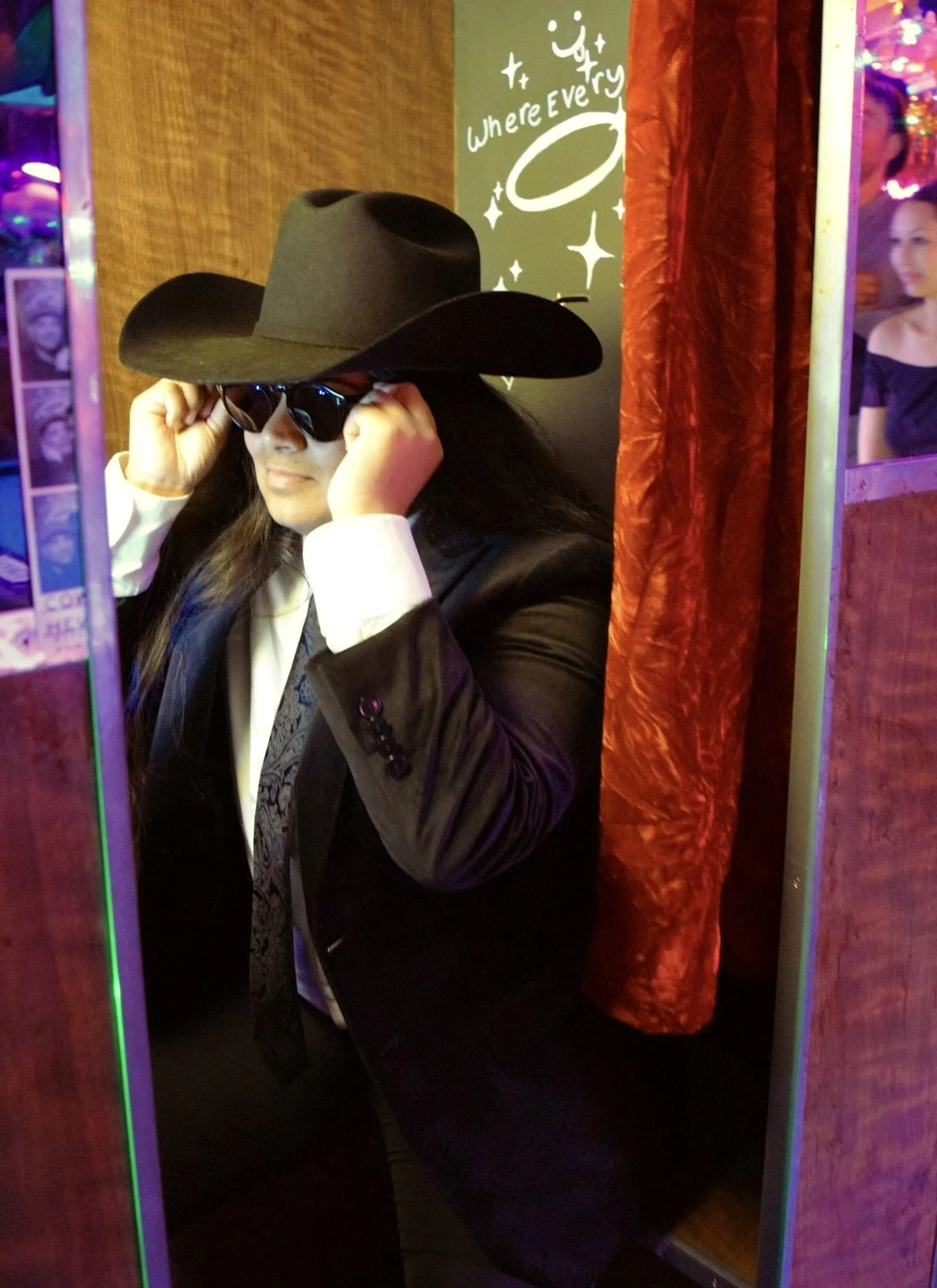 Person dressed in a black suit with a white shirt and patterned tie, wearing a large black cowboy hat and sunglasses, inside a photo booth with a red curtain and green wall with white text and design.