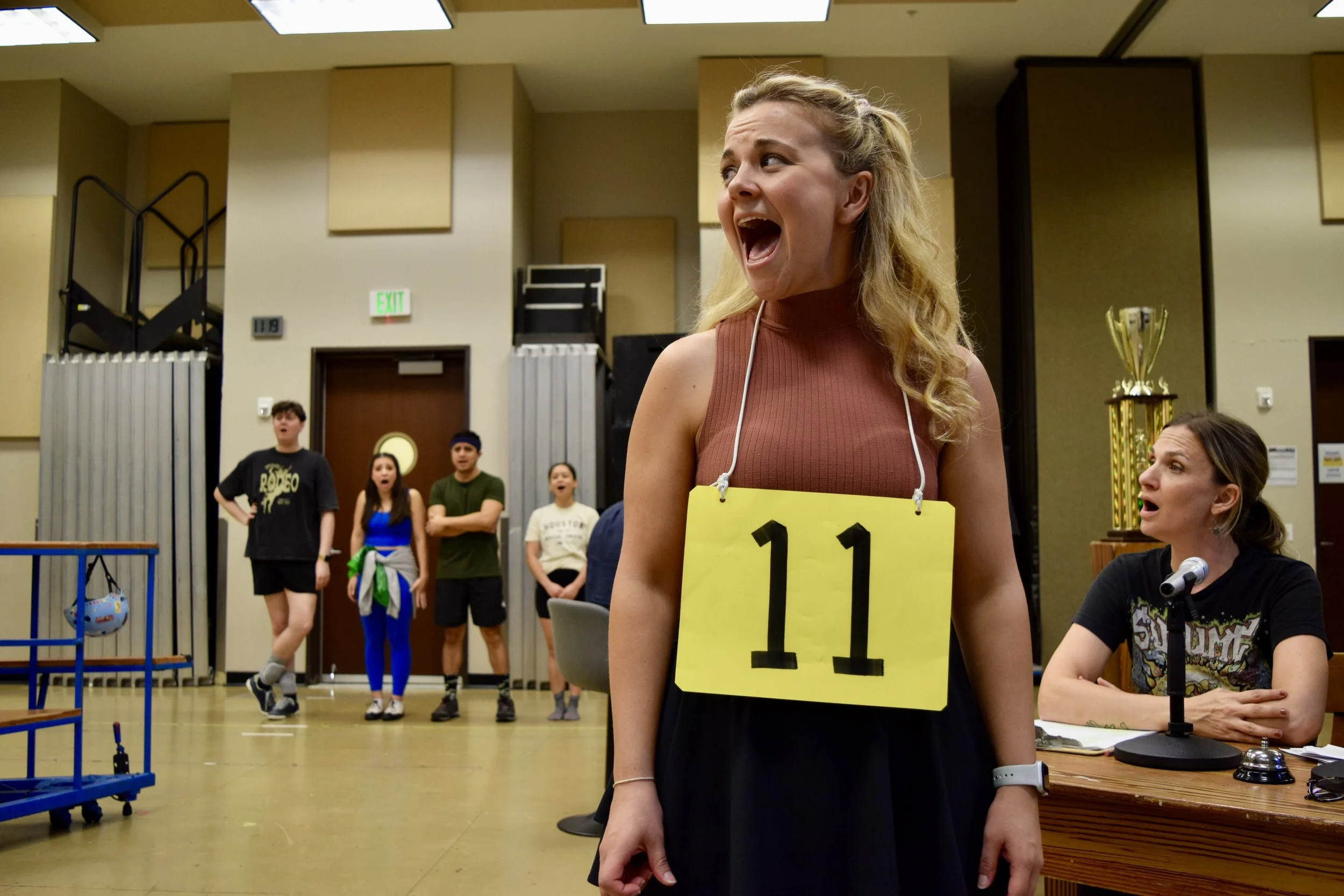 A woman wearing a brown sleeveless top and black skirt with a yellow audition number 11 around her neck, appears surprised or excited. She is in a large indoor space with several other people in the background, some watching and some standing with expressions of surprise or curiosity.