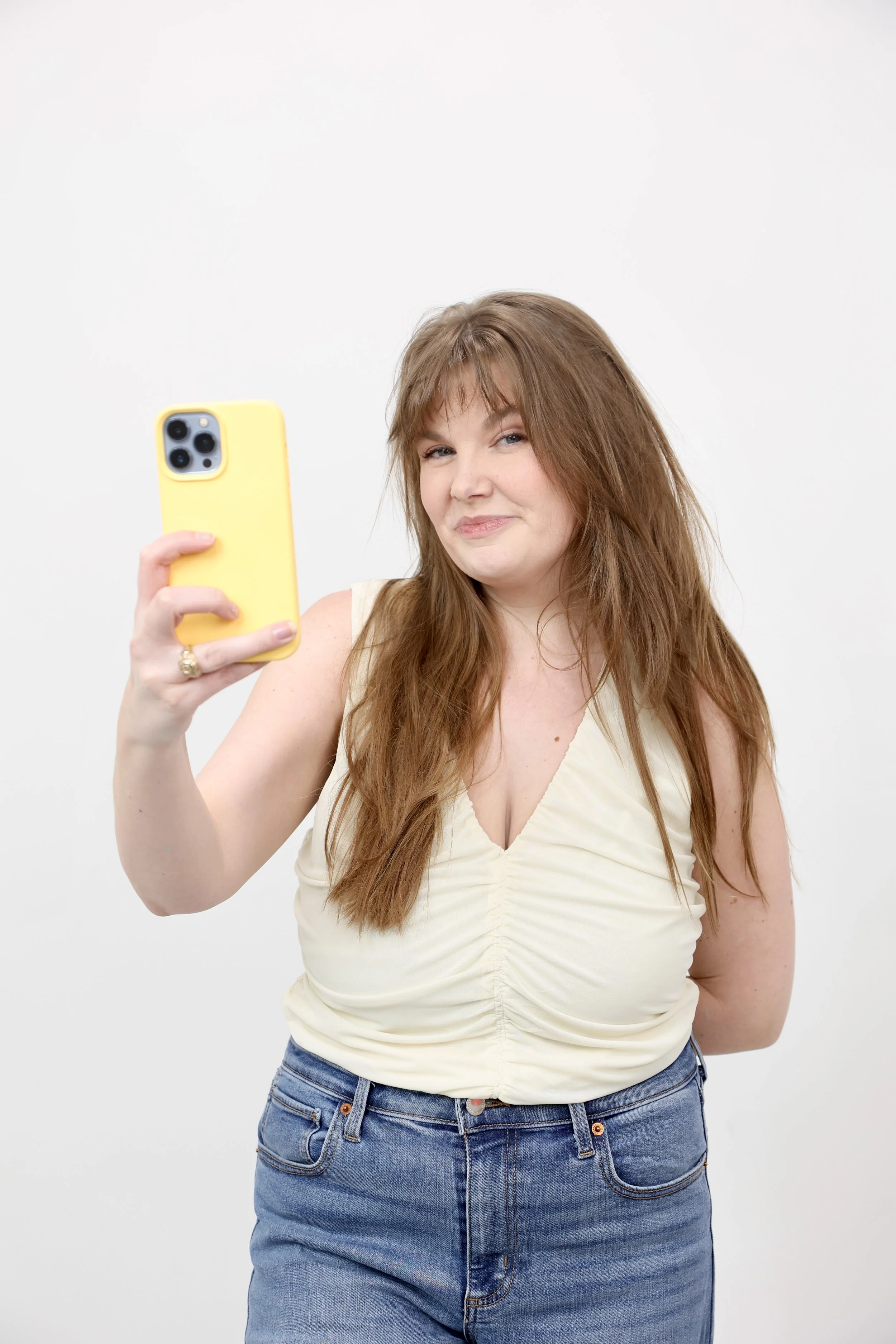 Woman with long reddish hair taking a selfie with a yellow phone case, smiling slightly, against a plain white background.