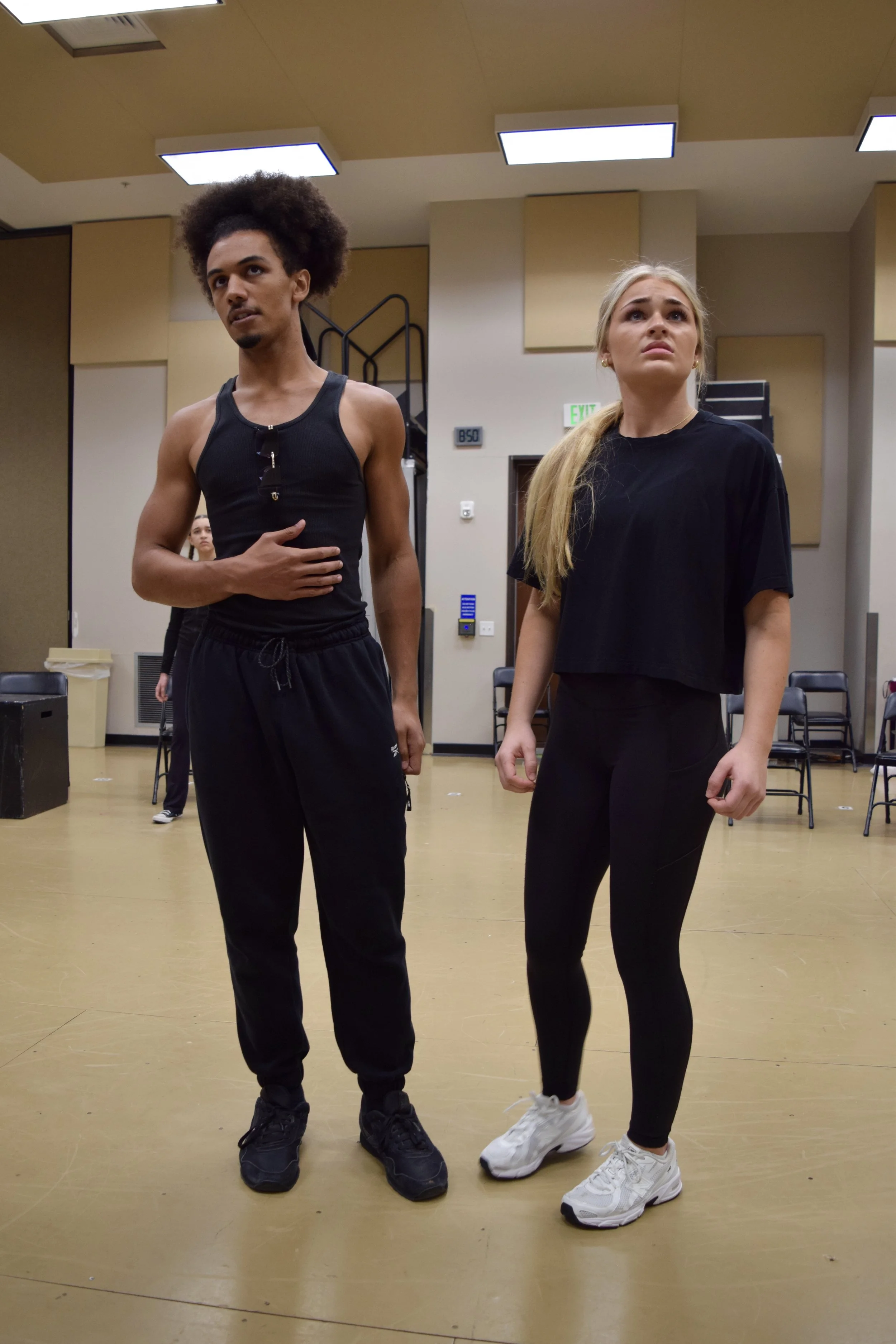 Two young adults, a man and a woman, standing in a dance studio or rehearsal space with chairs in the background. The man has curly hair, a sleeveless black shirt, and black pants. The woman has long blonde hair, a loose black top, and black leggings