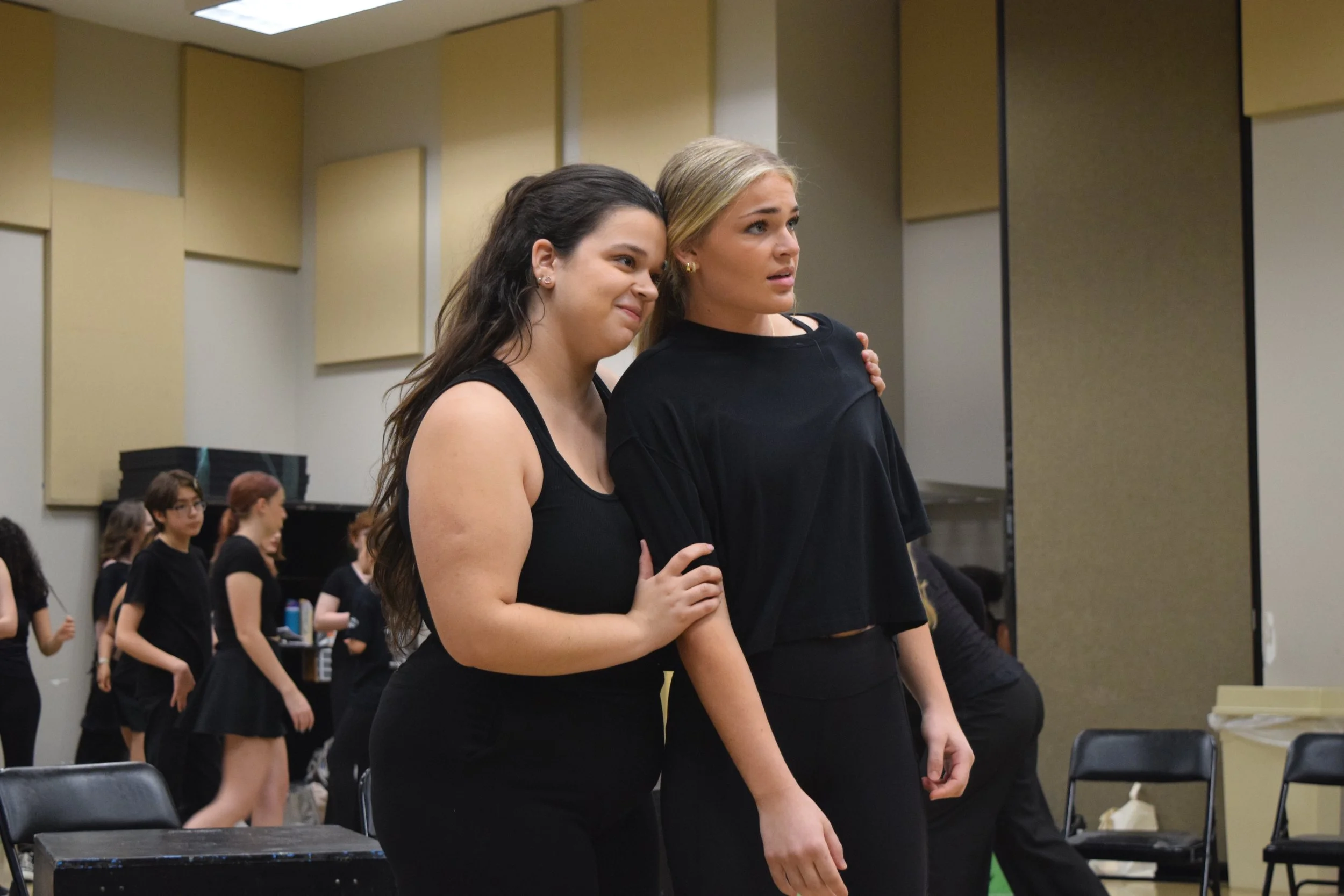 Two women stand closely together in a rehearsal room, with other women in black clothing in the background, possibly preparing for a performance.
