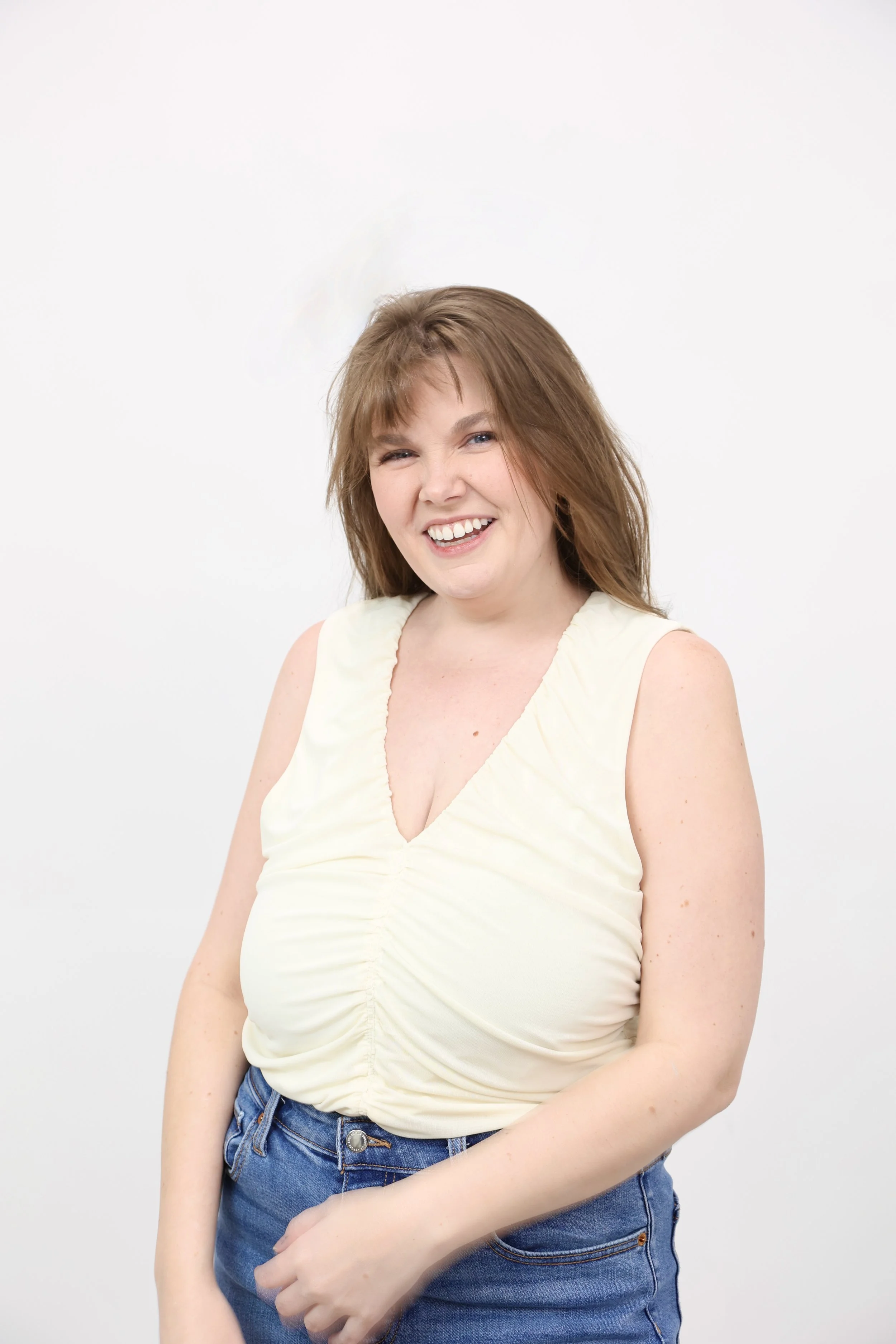 A young woman with light brown hair, smiling and winking, standing against a white background, wearing a sleeveless cream-colored top and blue jeans.