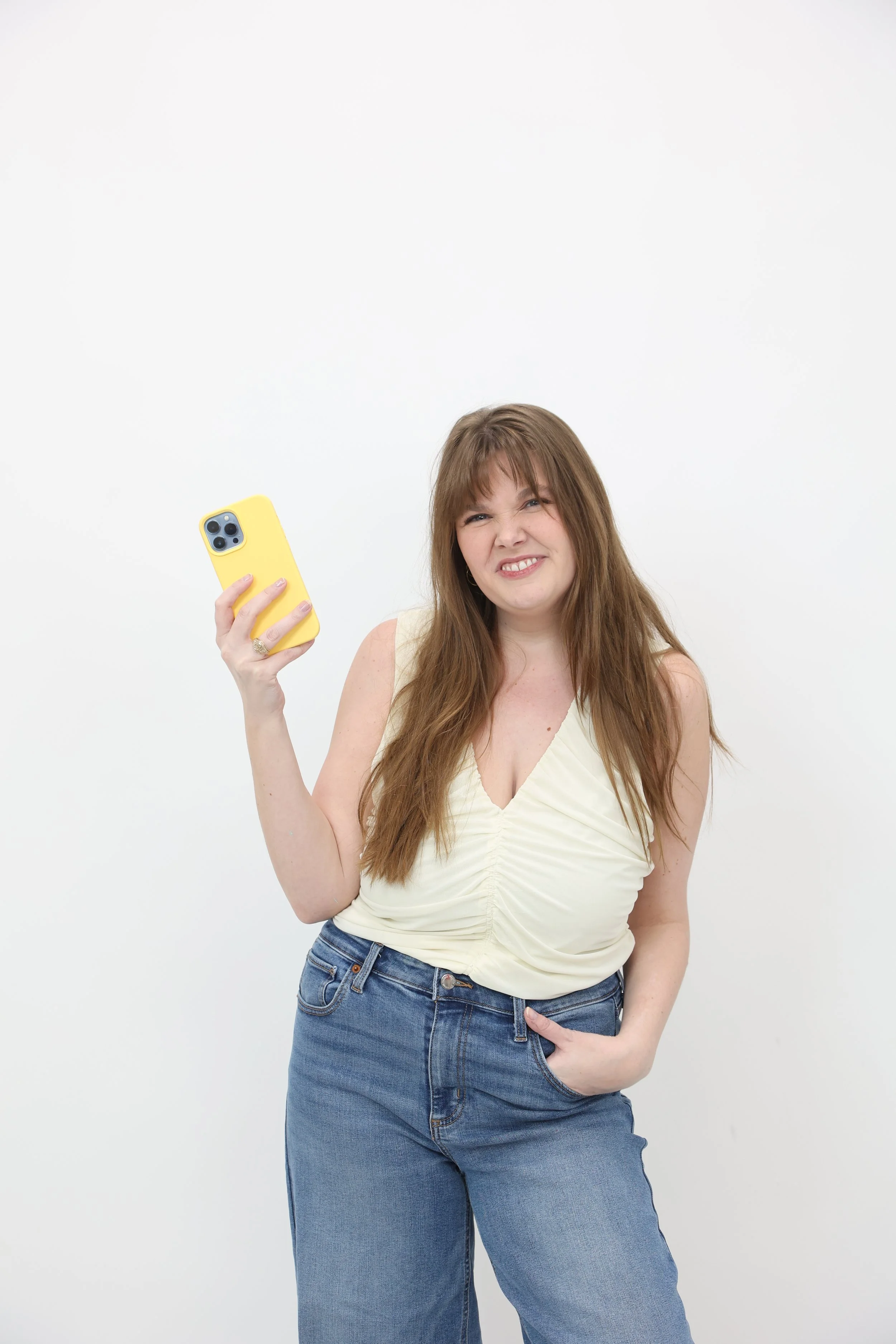 A woman with long brown hair making a grimace while holding a yellow smartphone in her right hand and placing her left hand in her pocket against a plain white wall.