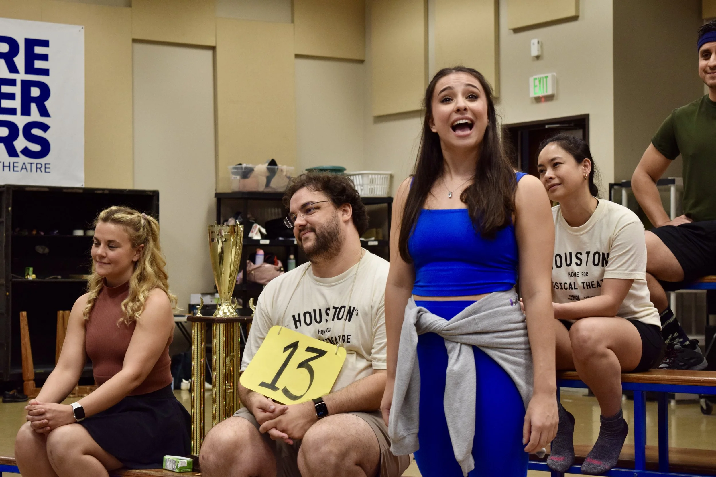 Group of people sitting and standing on a court, likely during a theatrical or dance rehearsal, with a woman in a blue dress speaking and others listening.