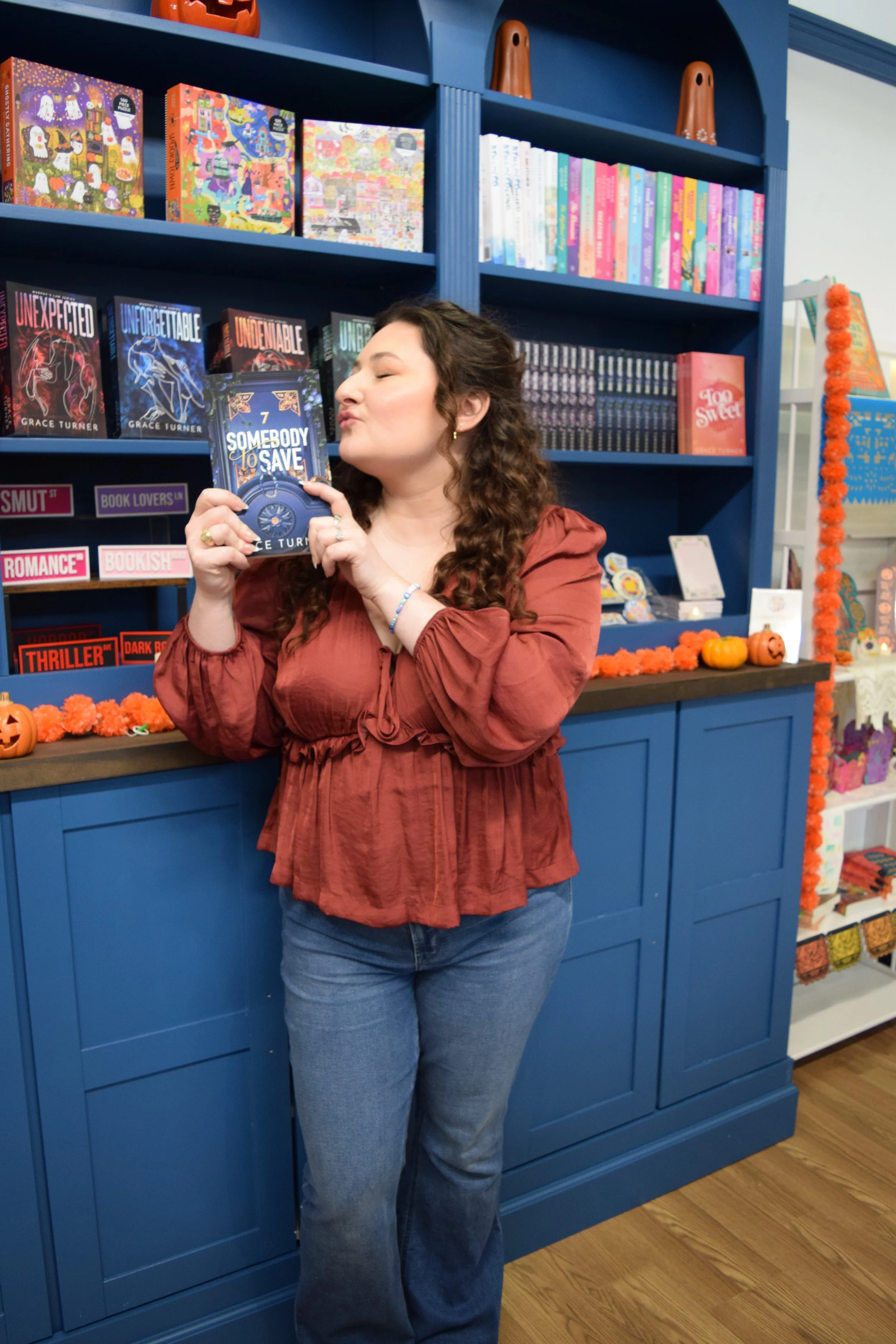 A woman with curly hair and wearing an orange blouse holding a book titled 'Somebody to Love' while standing in a bookstore decorated for Halloween with pumpkins and orange garland.