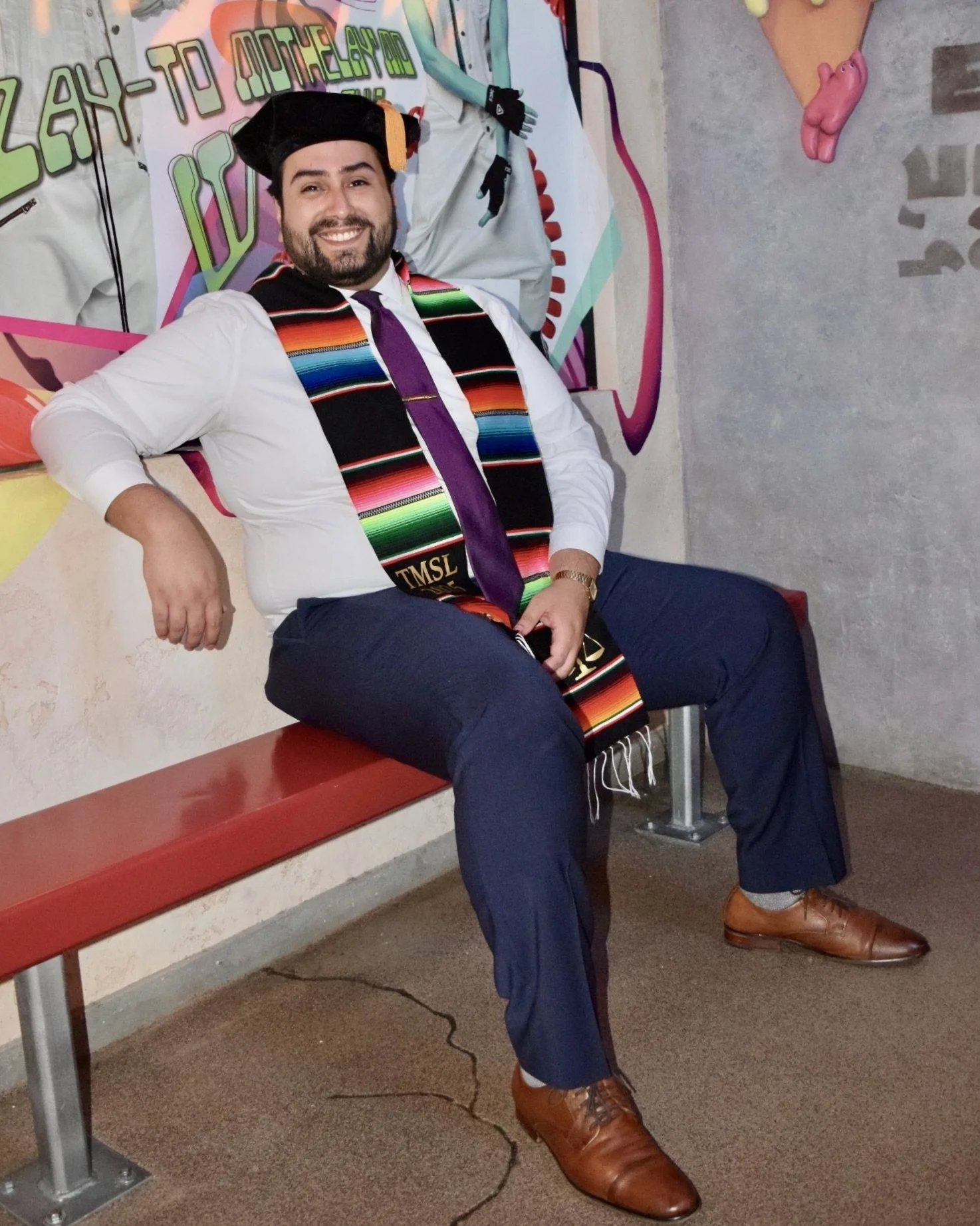 A man in a graduation cap and gown with a colorful sash, sitting on a red bench, smiling, in front of a mural with bright colors and designs.