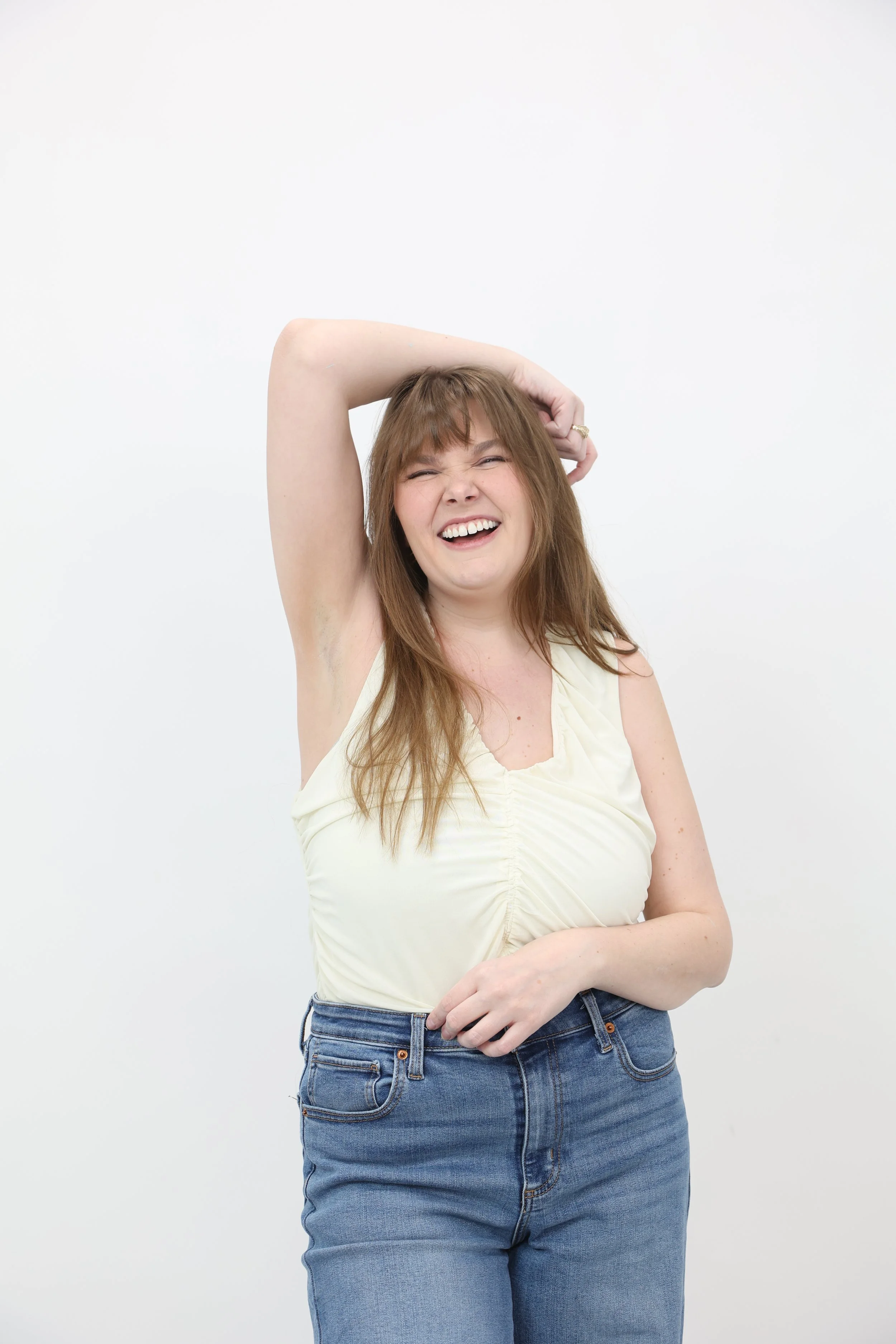 A young woman with long brown hair and fair skin laughing and posing with her right arm raised, in front of a plain white background.