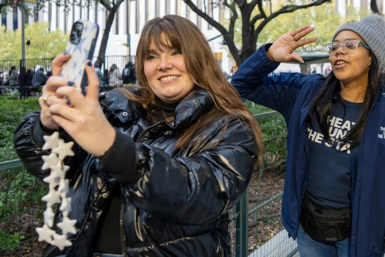 Two women taking a selfie outdoors in a park with trees and a fence in the background. One woman has long brown hair and is smiling while holding the phone, and the other woman, wearing glasses and a gray beanie, is making a gesture with her hand.