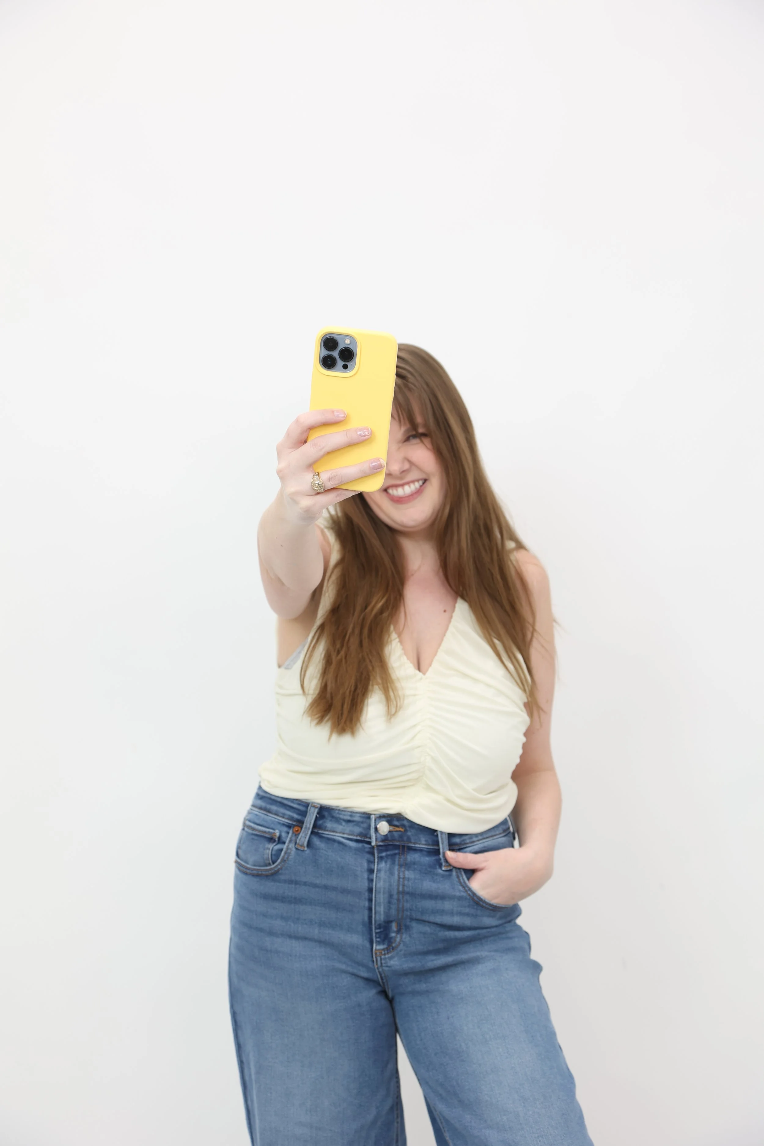 A woman with long, wavy brown hair taking a selfie with a yellow smartphone. She is smiling and wearing a light-colored, ruched sleeveless top and blue jeans against a plain white background.
